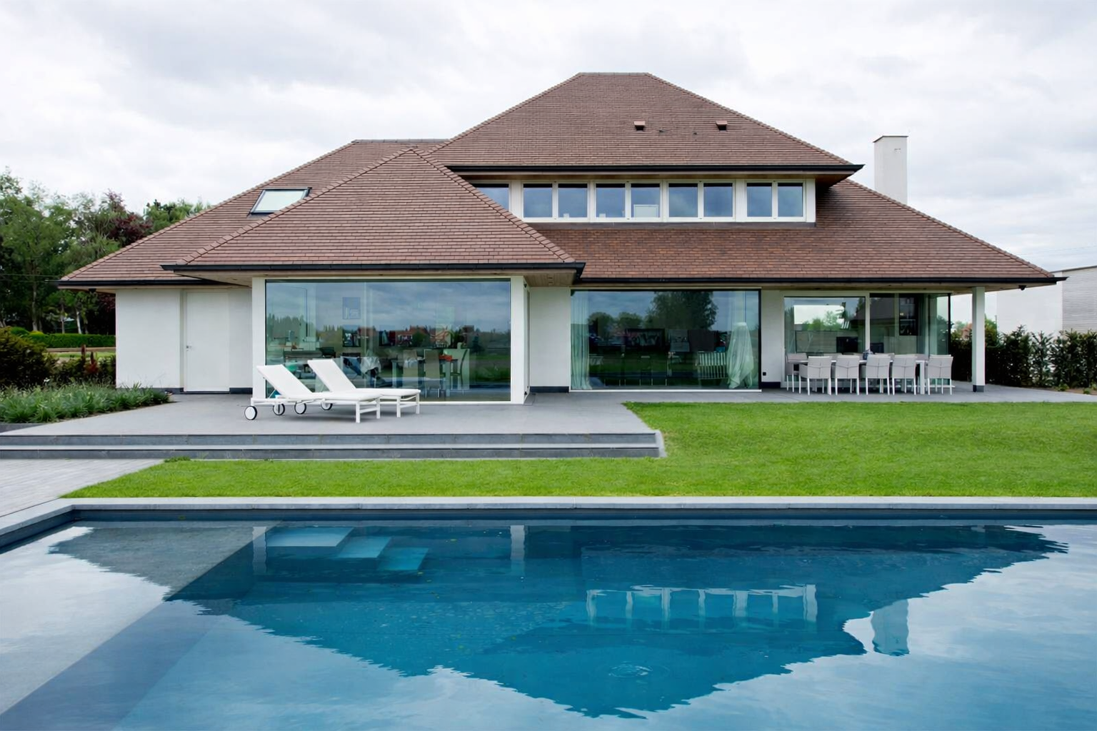 Modern house with large glass windows, a swimming pool, green lawn, outdoor lounge chairs, and a dining area, under a cloudy sky.