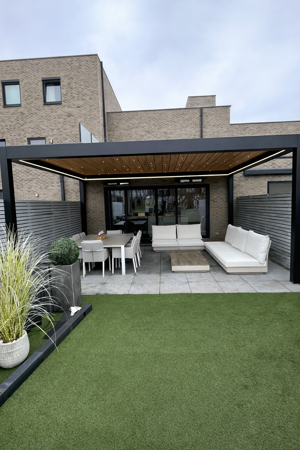 Modern outdoor patio area with white seating, dining table, potted plants, and a green lawn under a covered wooden roof, attached to a multi-story brick building.