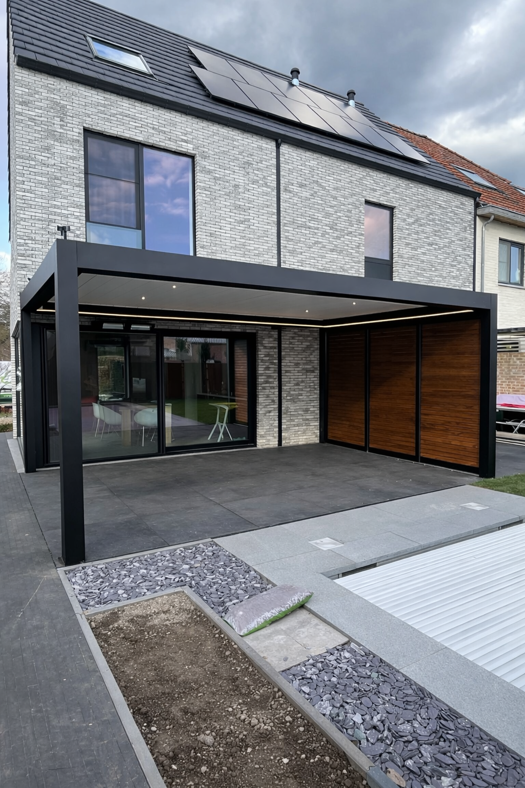 Modern house with gray brick exterior, black framed glass doors and windows, and solar panels on the roof. There is a covered patio area with a black frame and wooden panels, and some outdoor furniture visible through the glass doors. The yard in fro
