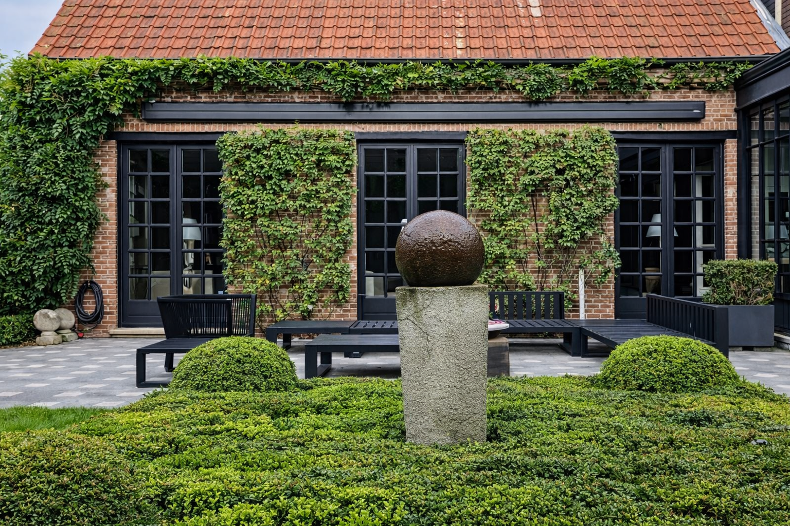 A modern backyard with a brick house, black-framed glass doors, and green climbing plants. There are two black benches and a stone pedestal with a round sculpture. Well-manicured bushes and a patterned patio are in the foreground.