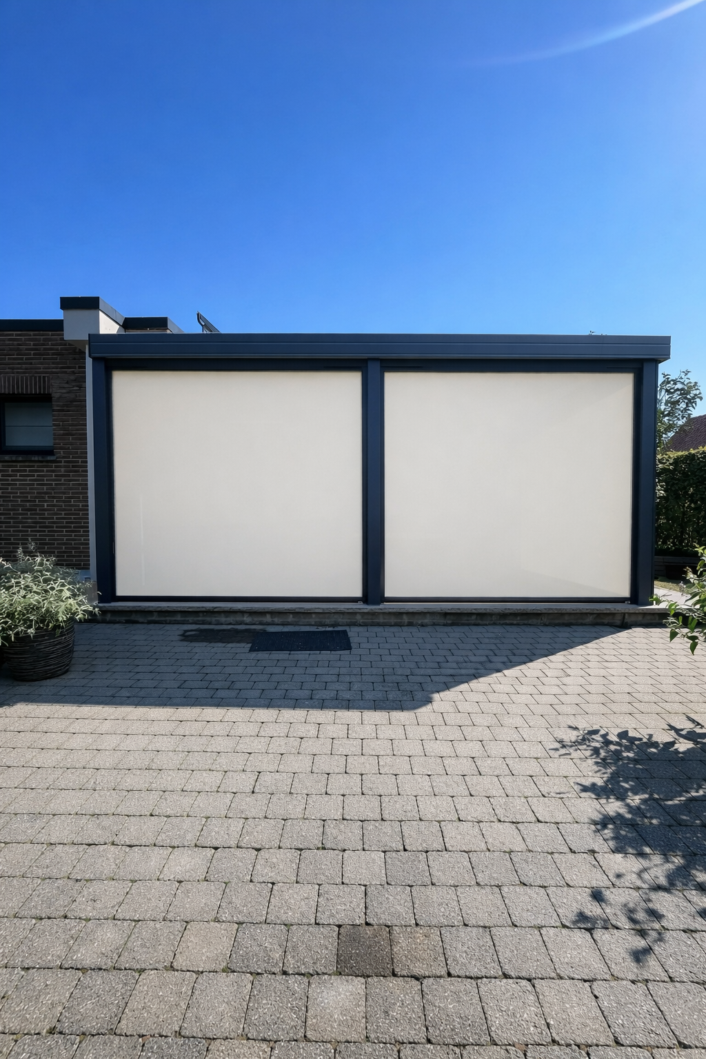 A modern building with large white panels on its exterior, situated on a paved courtyard with a plant in a pot to the left and shrubs on the right, under a clear blue sky.