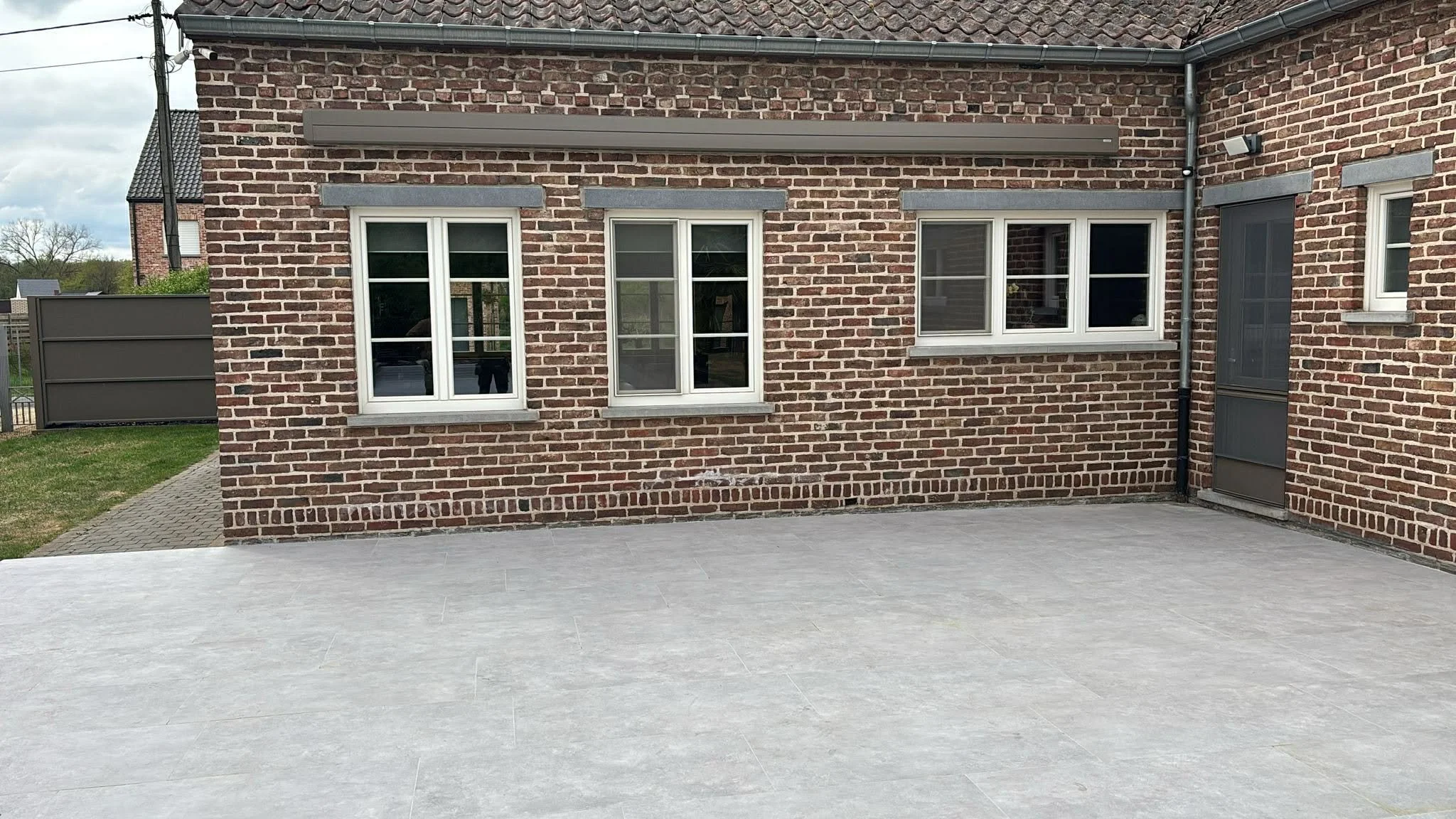 A brick house exterior with three white-framed double-hung windows, a screened door, gray trim, and an outdoor tiled patio area.