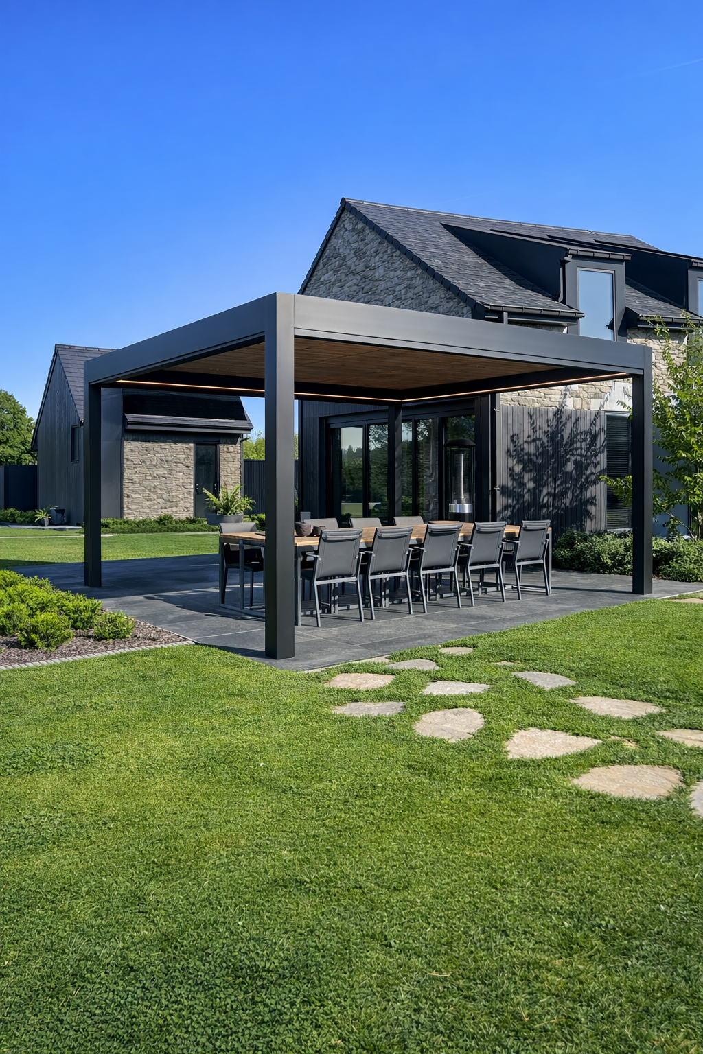 Modern outdoor dining area with a large table and ten chairs under a black metal pergola on a stone patio, adjacent to a contemporary house with dark exterior walls and windows, with a well-maintained green lawn and stepping stones in the yard.