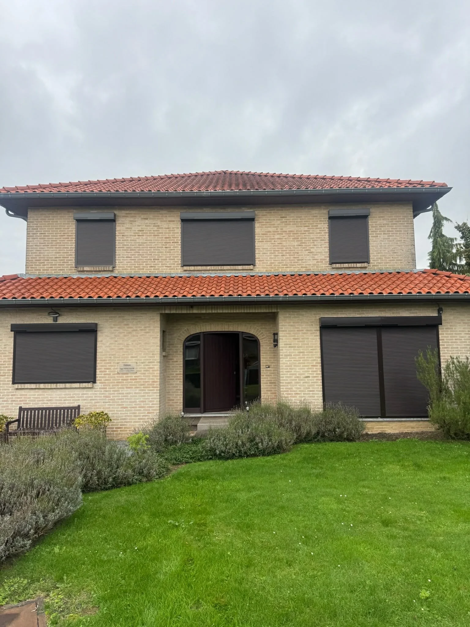 A two-story house with yellow brick walls, dark roller shutters on the windows, red tiled roof, and a dark front door, surrounded by green lawn and shrubs under a cloudy sky.
