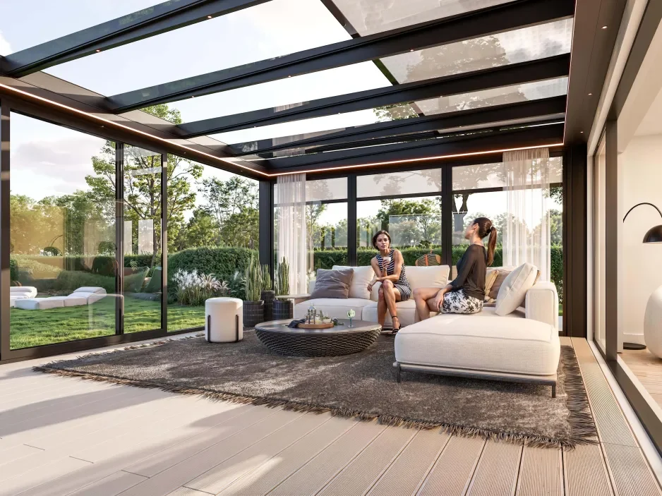 Two women sitting and talking on a modern white couch inside a glass-enclosed sunroom with a view of a lush green garden outside.