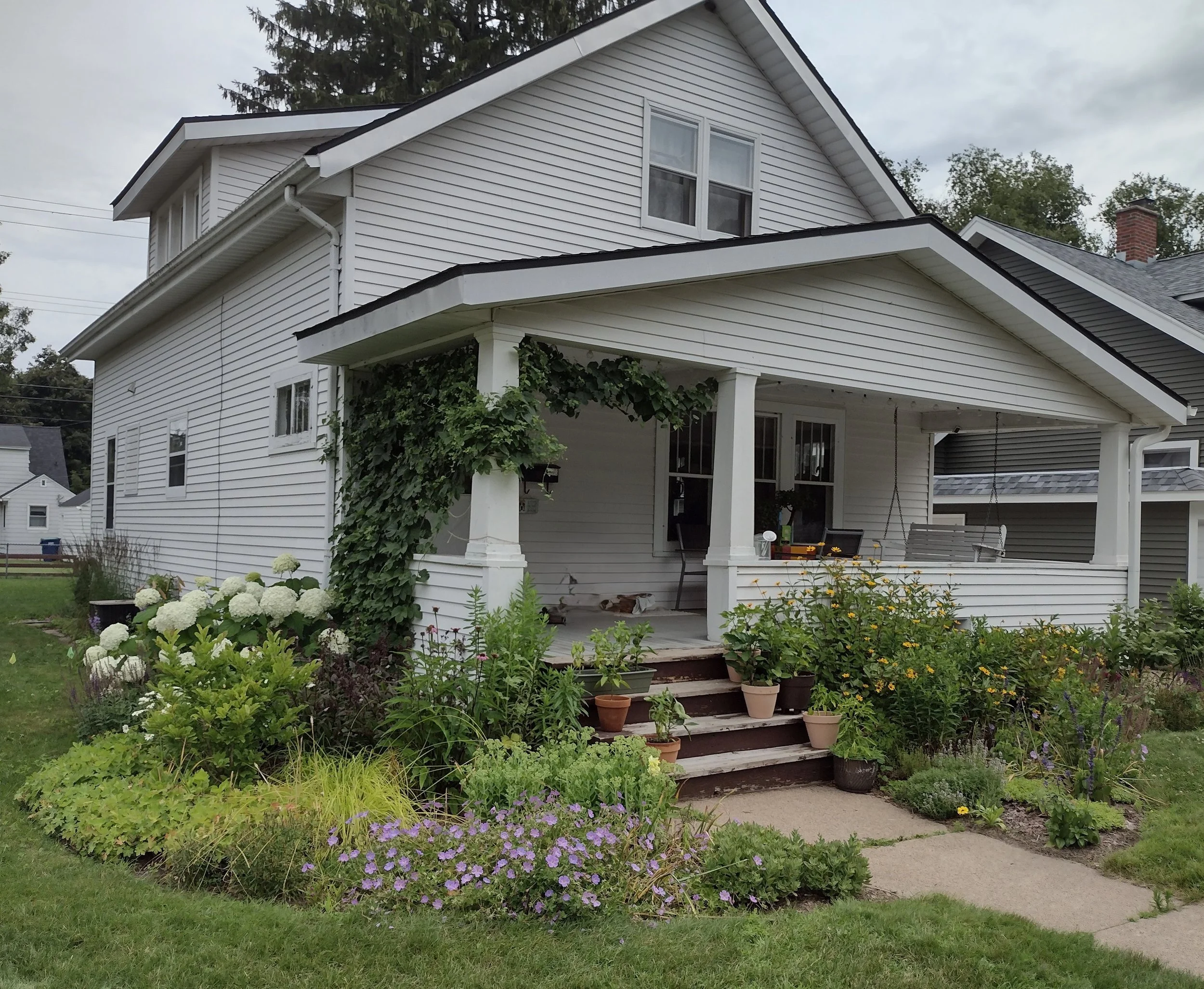 Front porch of a white house with a garden filled with various plants and flowers, some pots on the steps, and a swing hanging from the porch ceiling.