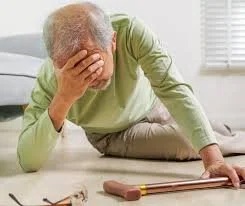 An elderly person sitting on the floor with a hand on their forehead, appearing distressed, in a room with sunlight and a window.