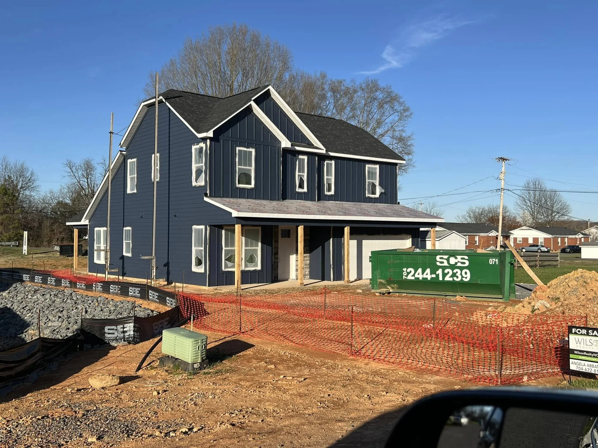 Under construction two-story blue house with black roofing, scaffolding, a green dumpster, and orange safety fencing.