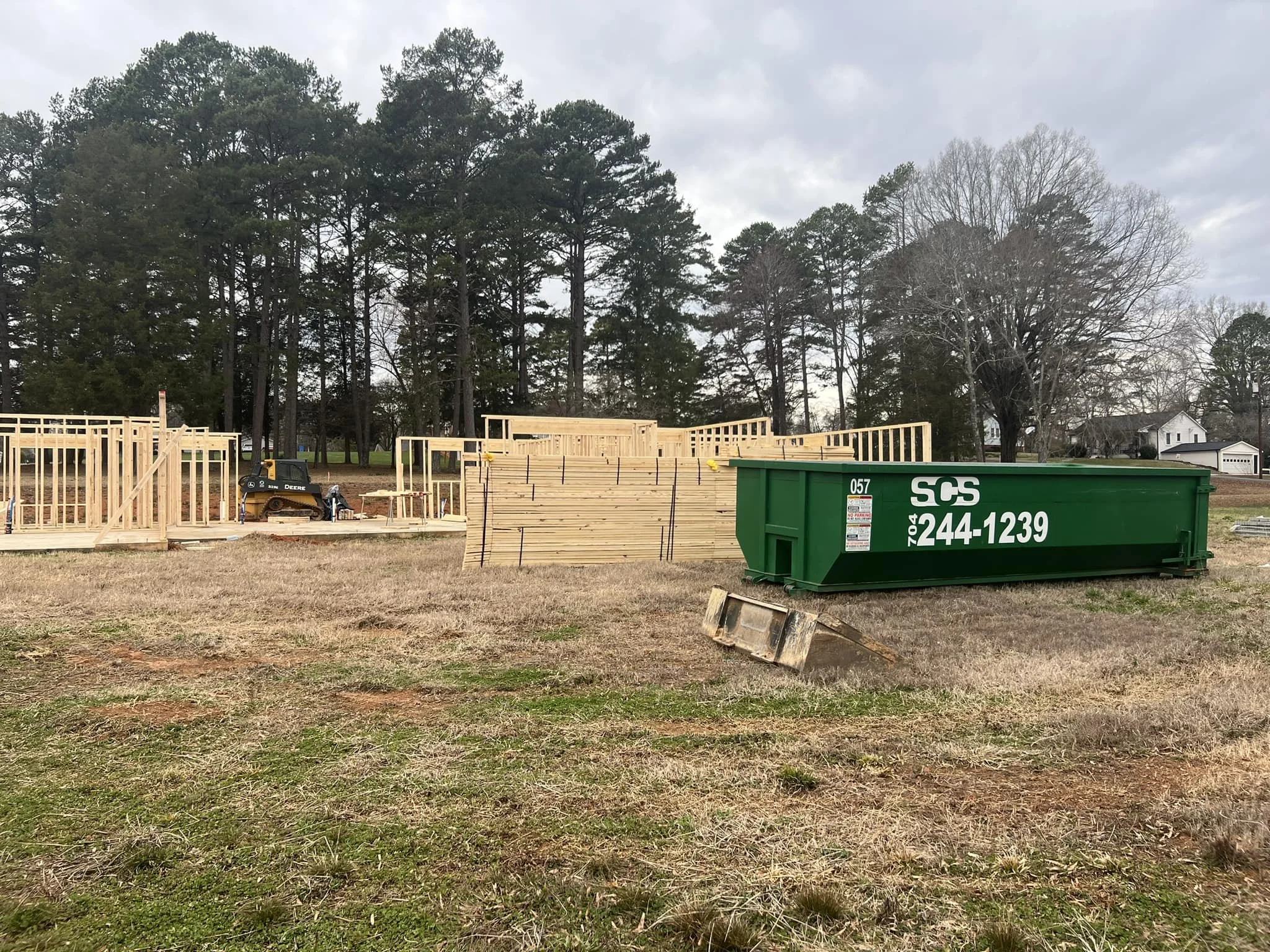 Construction site with wooden framing structures, a green waste bin, and construction equipment, with trees and houses in the background.