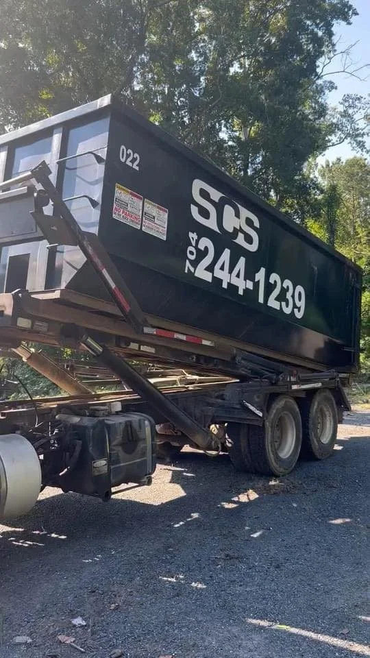 A large black dumpster trailer with white lettering marked 'SCS' and identification numbers, attached to a truck, parked outdoors on gravel with trees in the background.