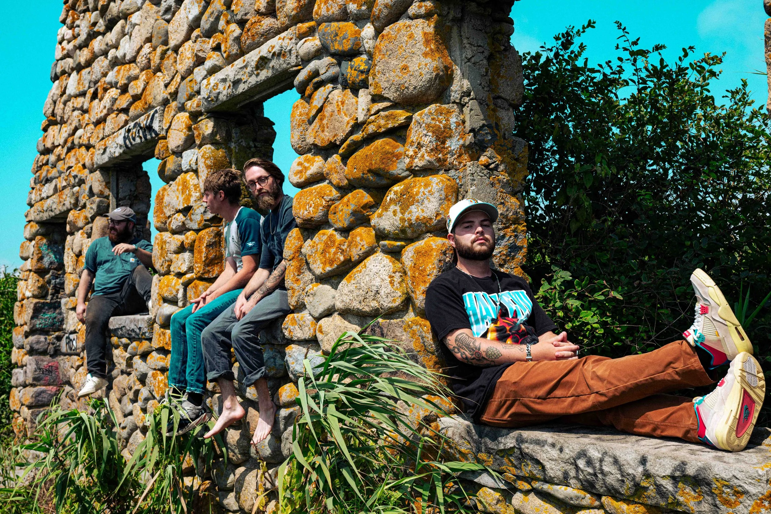 Five men sitting on a stone wall with some greenery around it. The men are relaxing, with some sitting and one lying down, variously dressed in casual clothing and sneakers or barefoot, under a bright blue sky.