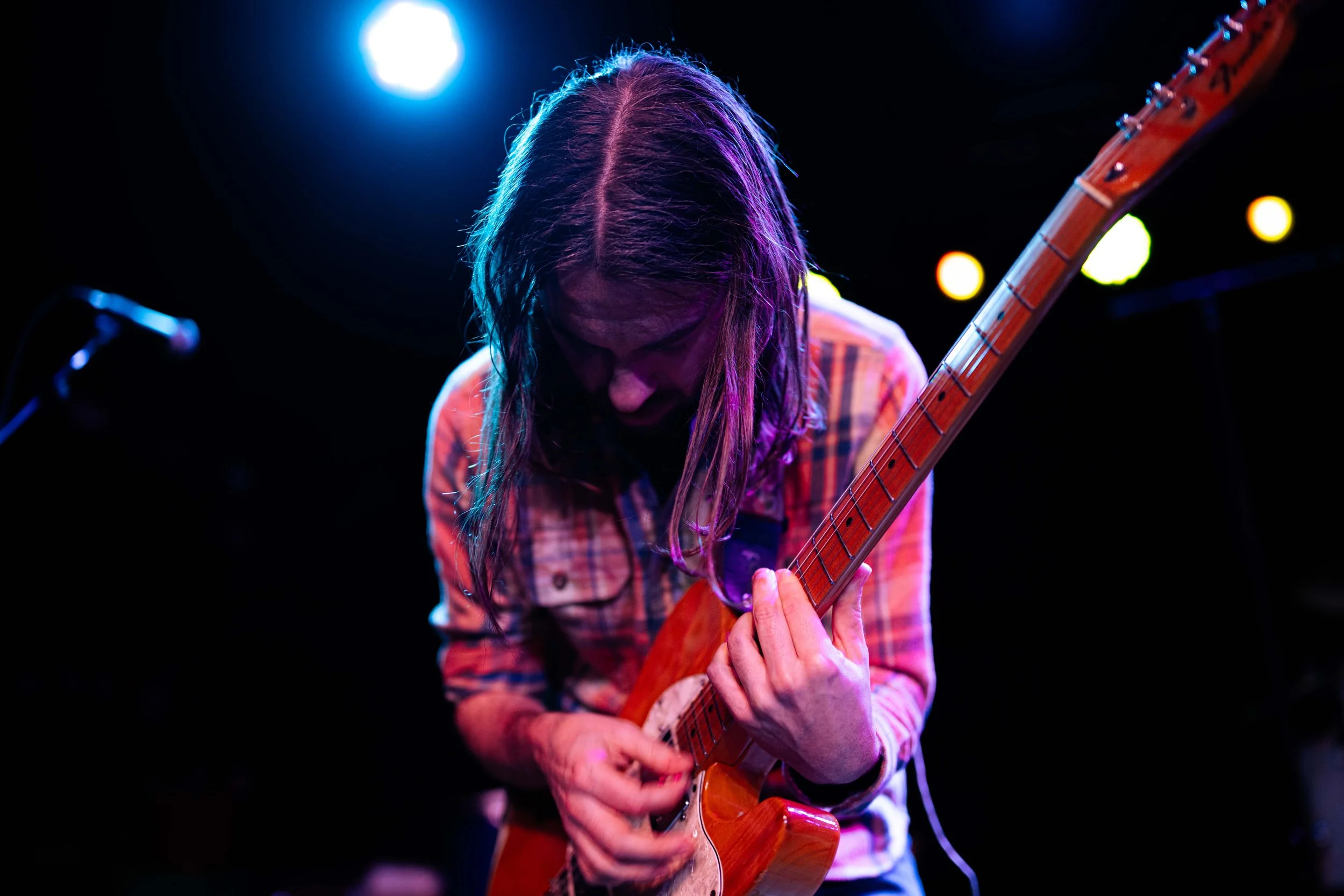 A person with long hair wearing a plaid shirt playing an electric guitar on a stage with colorful lighting.