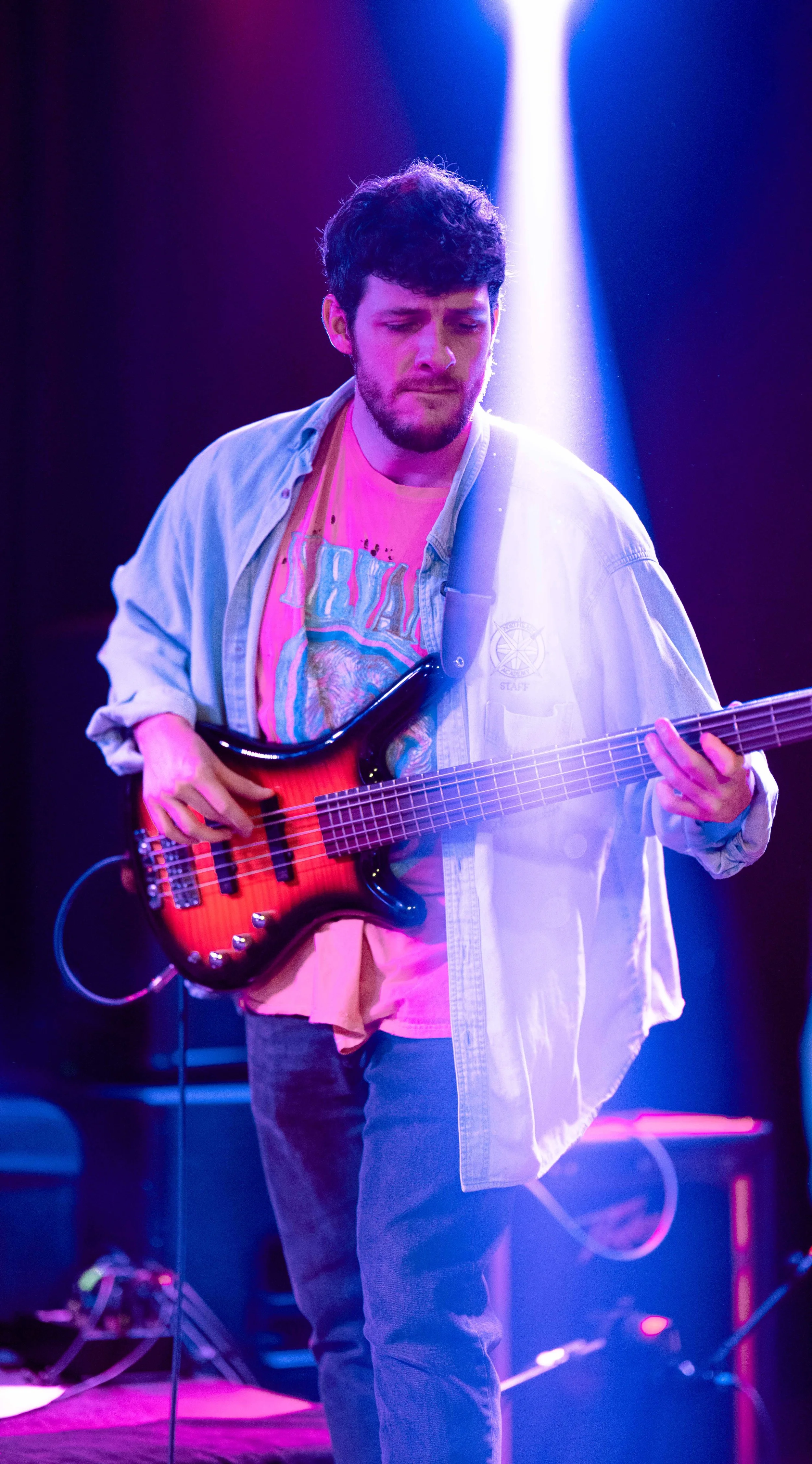 A young man with dark, curly hair and a beard playing an electric bass guitar on stage under purple and blue lighting.