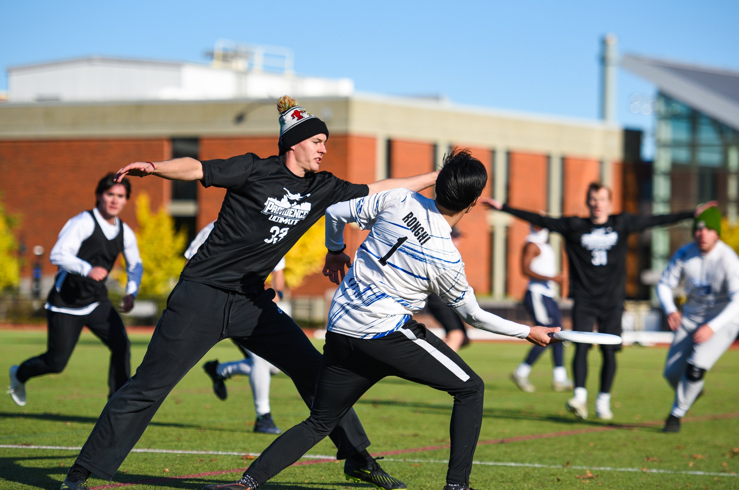 People playing ultimate frisbee on a field during daytime with a modern building and trees in the background.