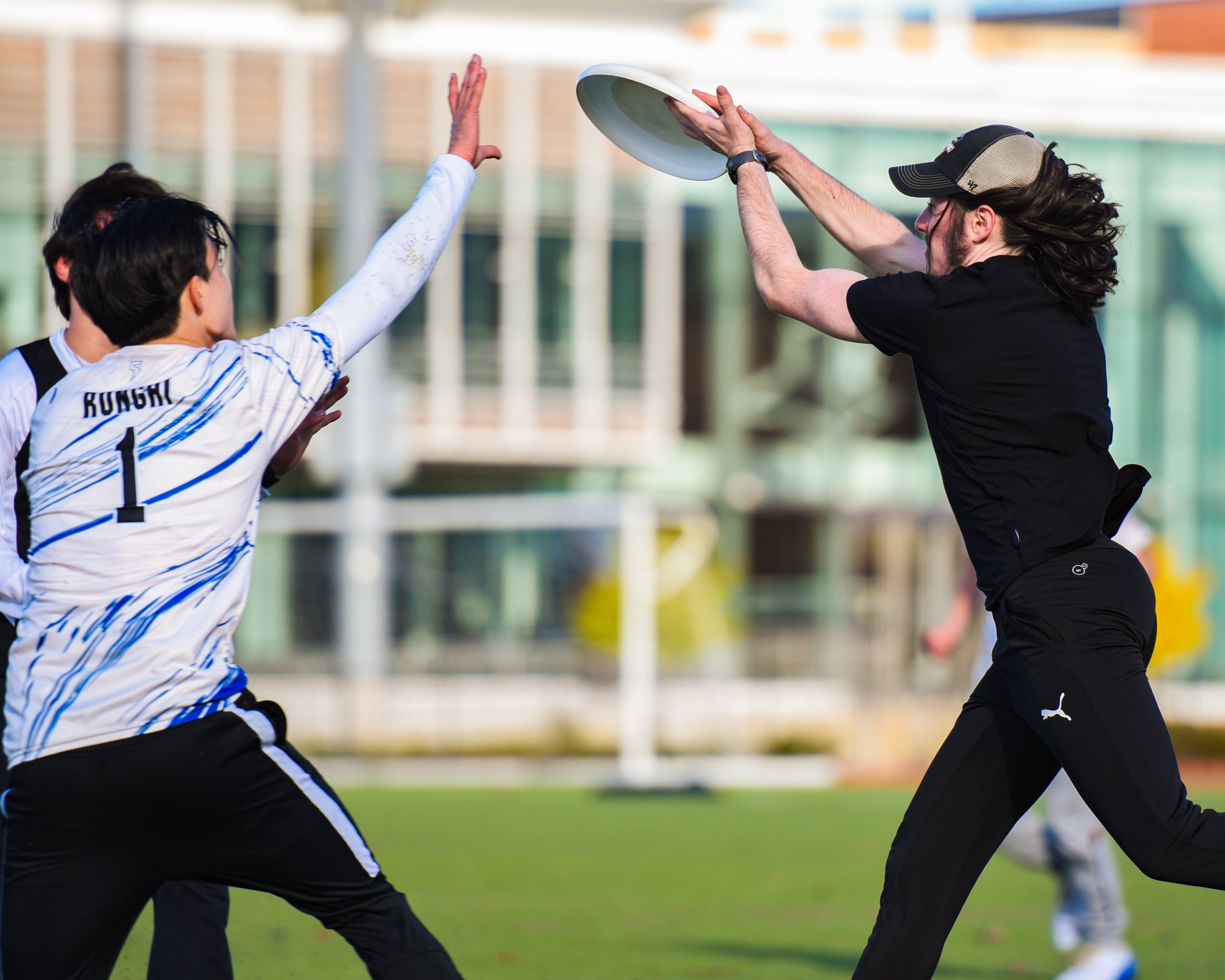 A man with long hair and a cap throws a frisbee while playing catch with a young man in a sports jersey on a grassy field.
