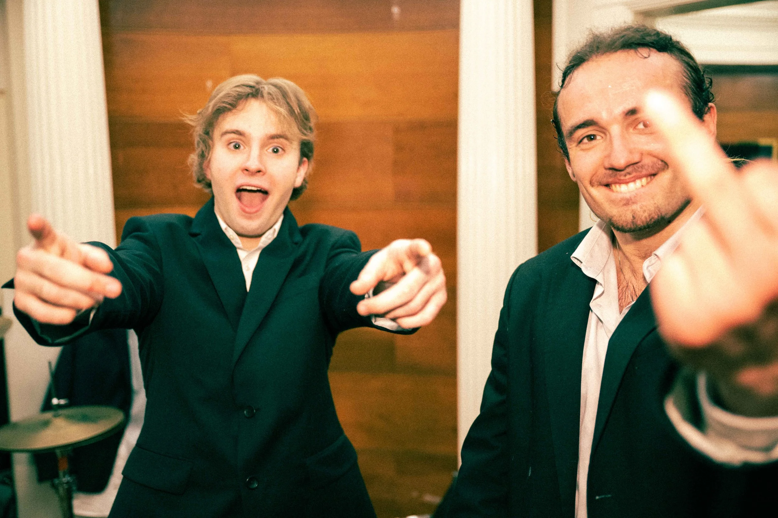 Two men in suits are smiling and pointing towards the camera at a social gathering.