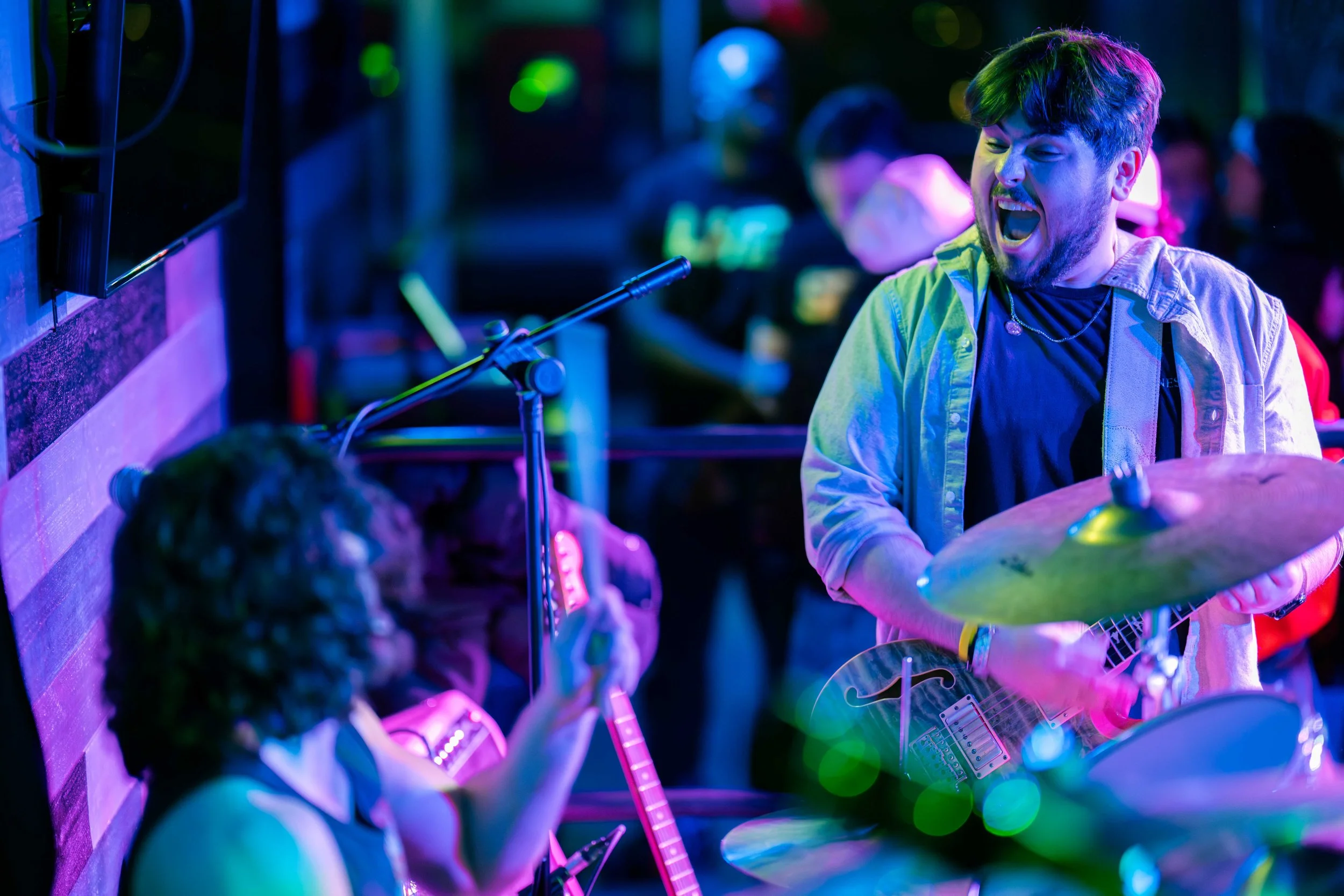 A lively setting with a young man playing guitar and singing passionately into a microphone, while a young girl with curly hair strums an acoustic guitar. The scene is vibrant with colorful lighting, and other people are visible in the background enj