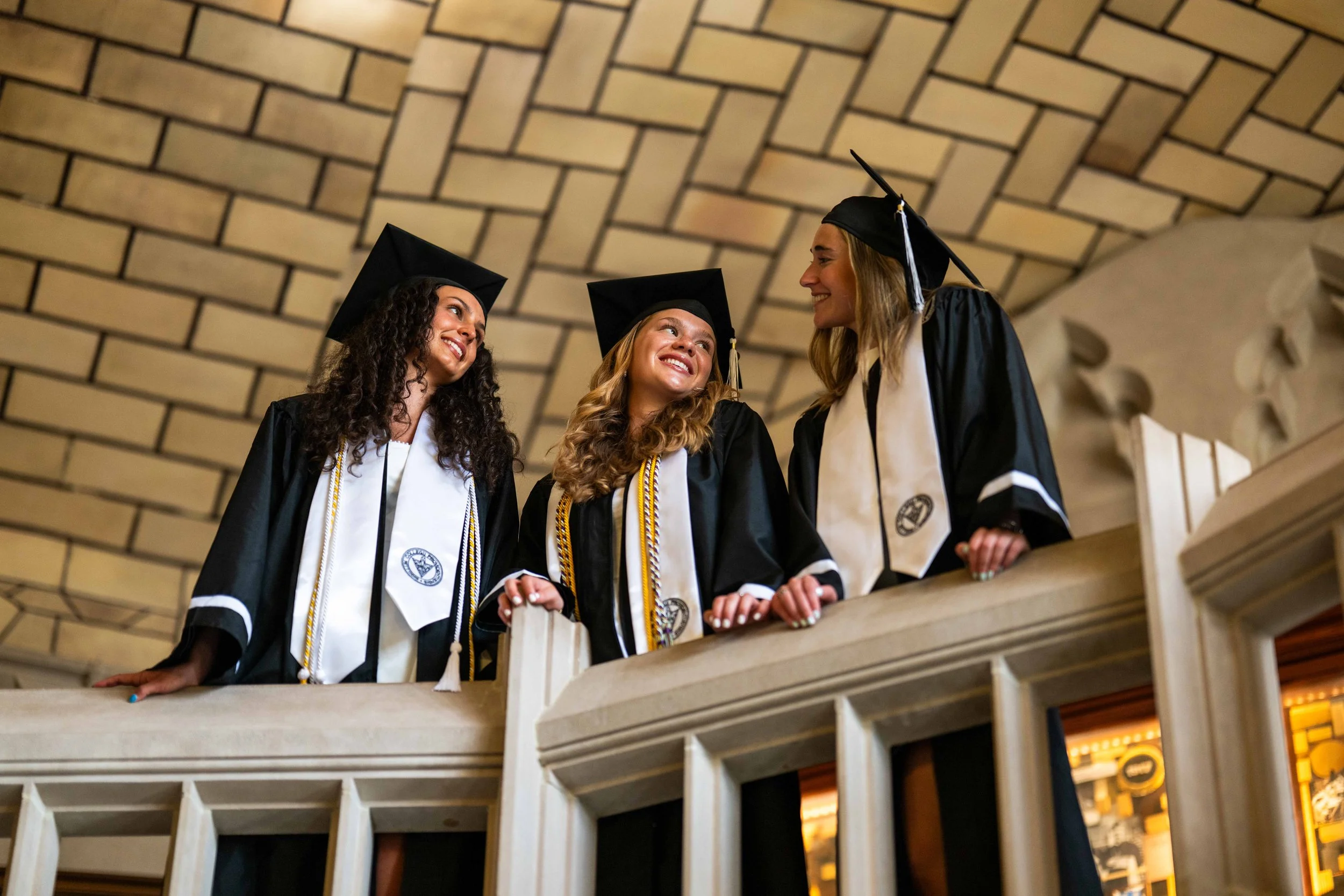 Three female graduates in caps and gowns smiling and talking to each other on a balcony with a stone ceiling.