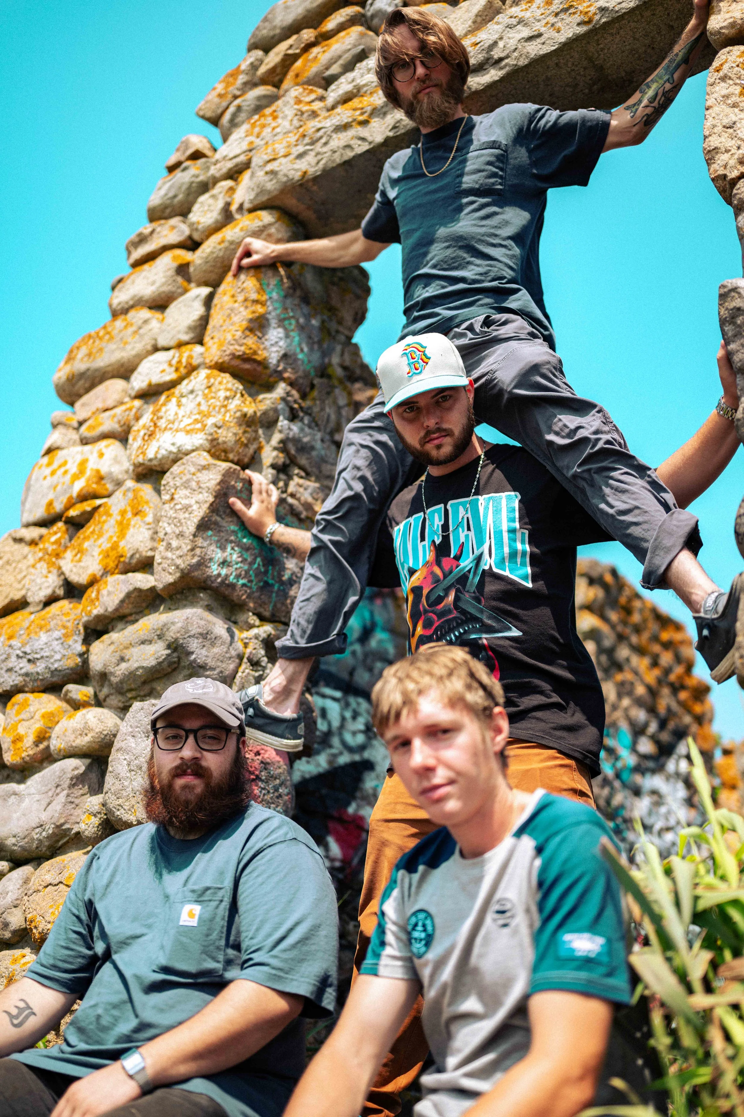 Four young men with casual clothing and tattoos in front of a stone structure with graffiti, under a clear blue sky.