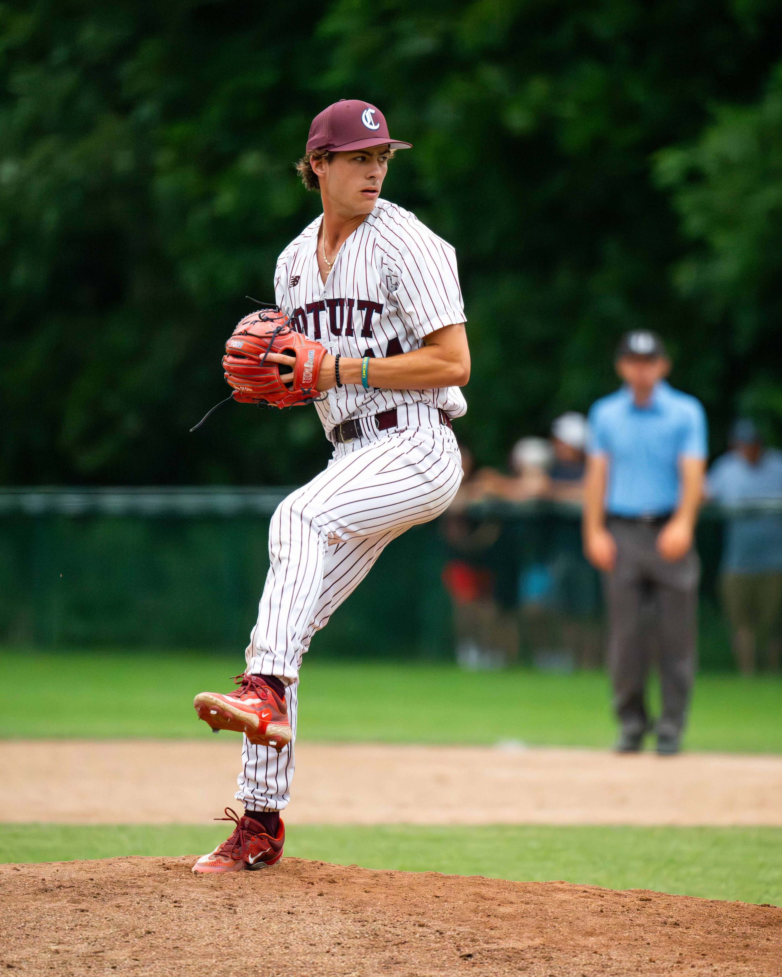 A young male baseball player in a white and maroon striped uniform and cap preparing to pitch on the mound during a game, with a blurred referee and spectators in the background.
