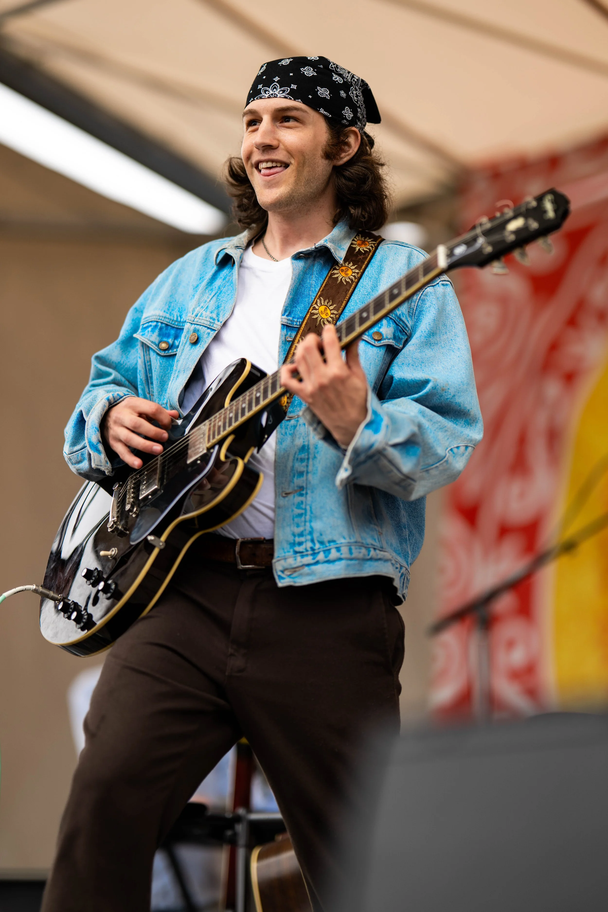 A singer-songwriter performing with an electric guitar on stage, wearing a denim jacket, a white t-shirt, brown pants, and a black bandana, smiling while playing.