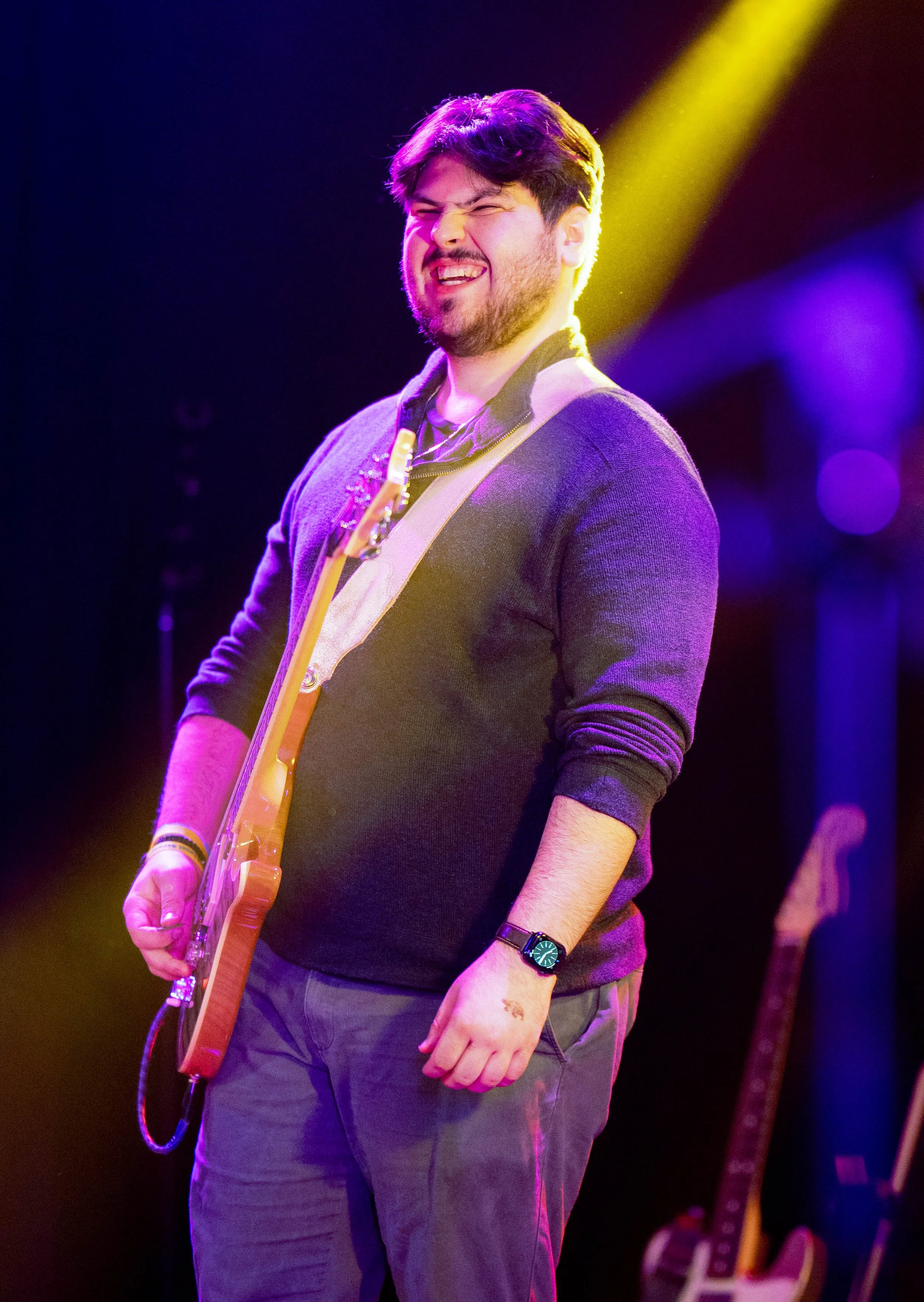 A young man with dark hair and a beard smiling and playing an electric guitar on stage with colorful purple and yellow lighting in the background.