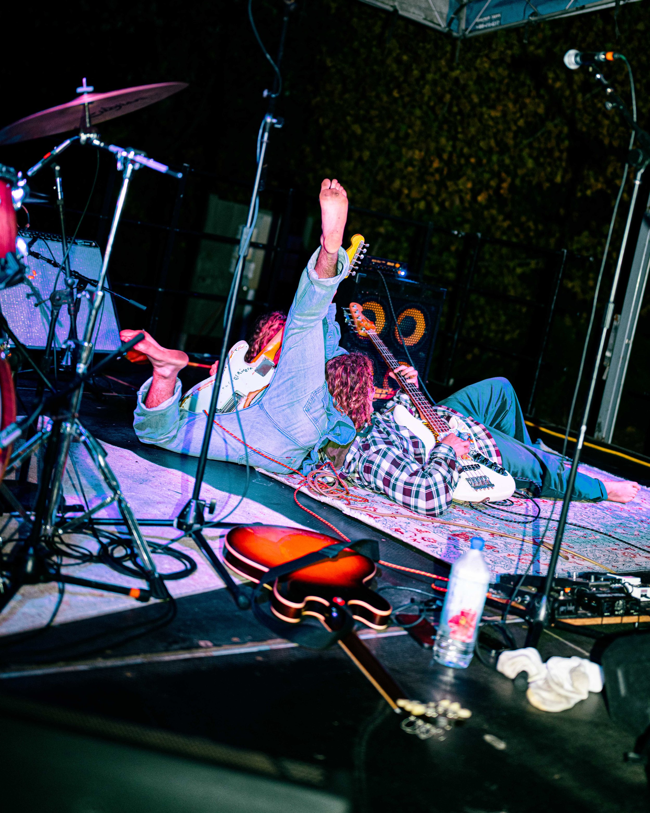 Two musicians lying on the stage floor with guitars, surrounded by microphones, cables, and equipment, during a live music performance.