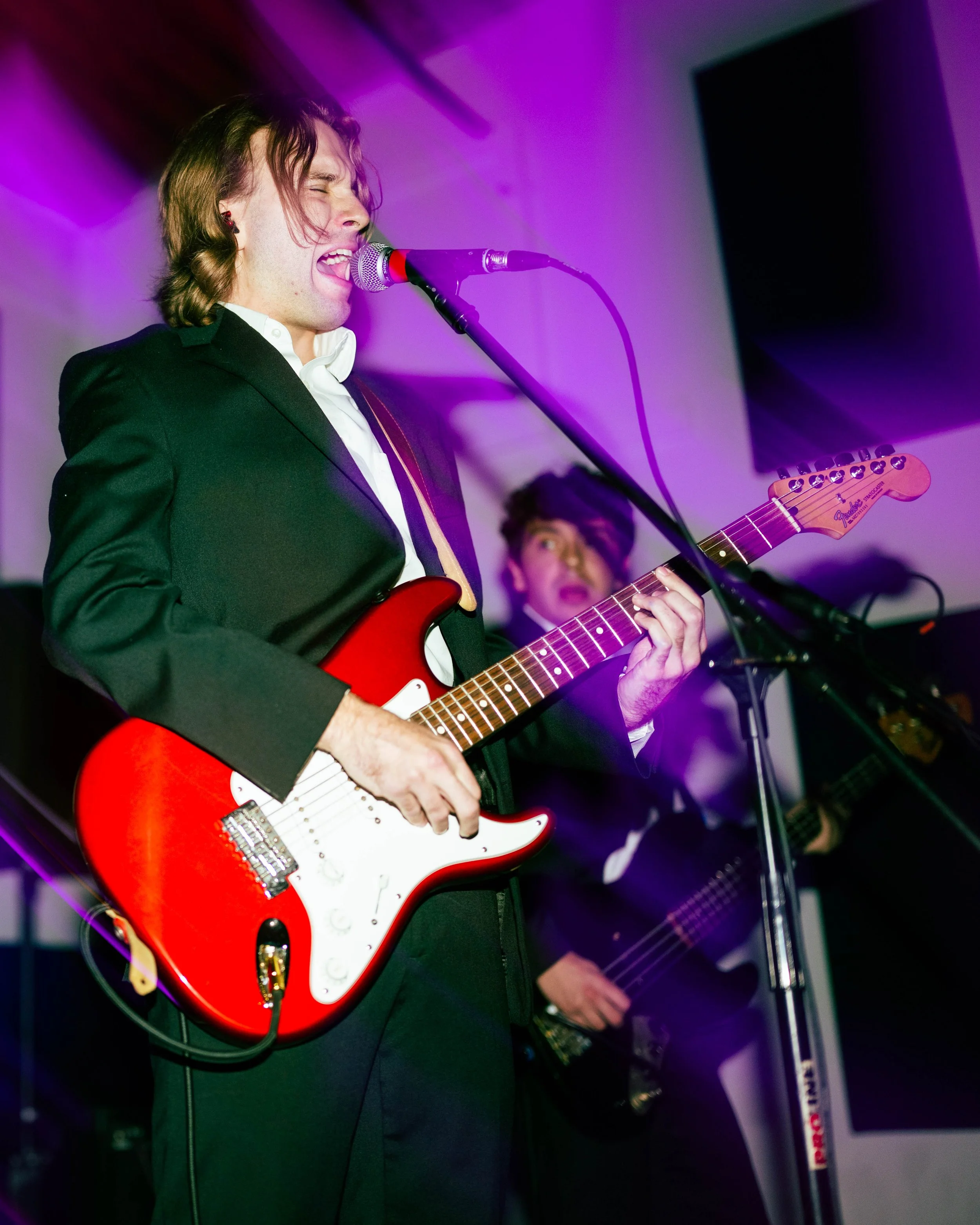 A man in a black suit jacket and white shirt playing a red electric guitar and singing into a microphone during a performance, with another musician in the background playing a bass guitar.