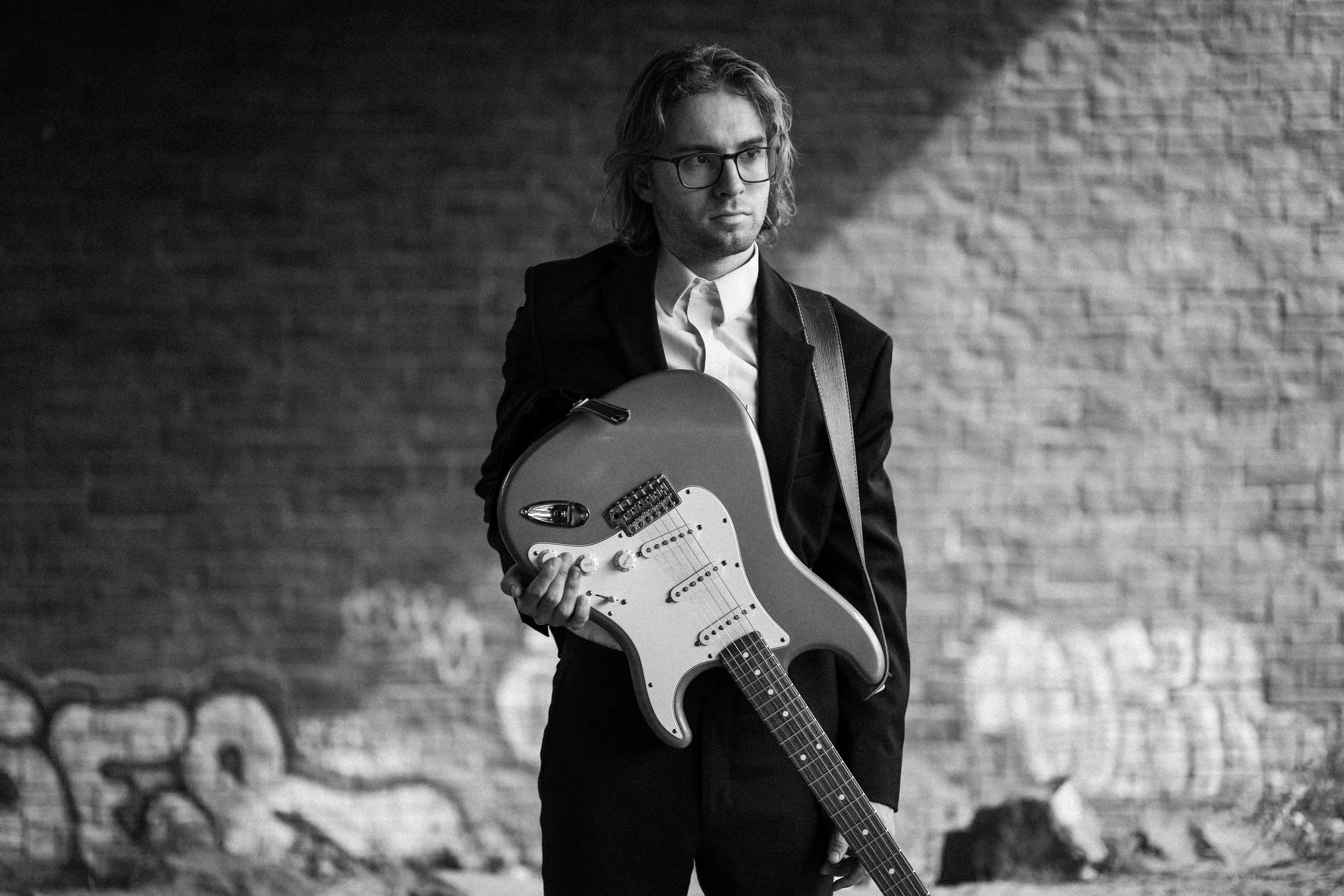A man with glasses and long hair in a suit holding an electric guitar in a black-and-white photo.