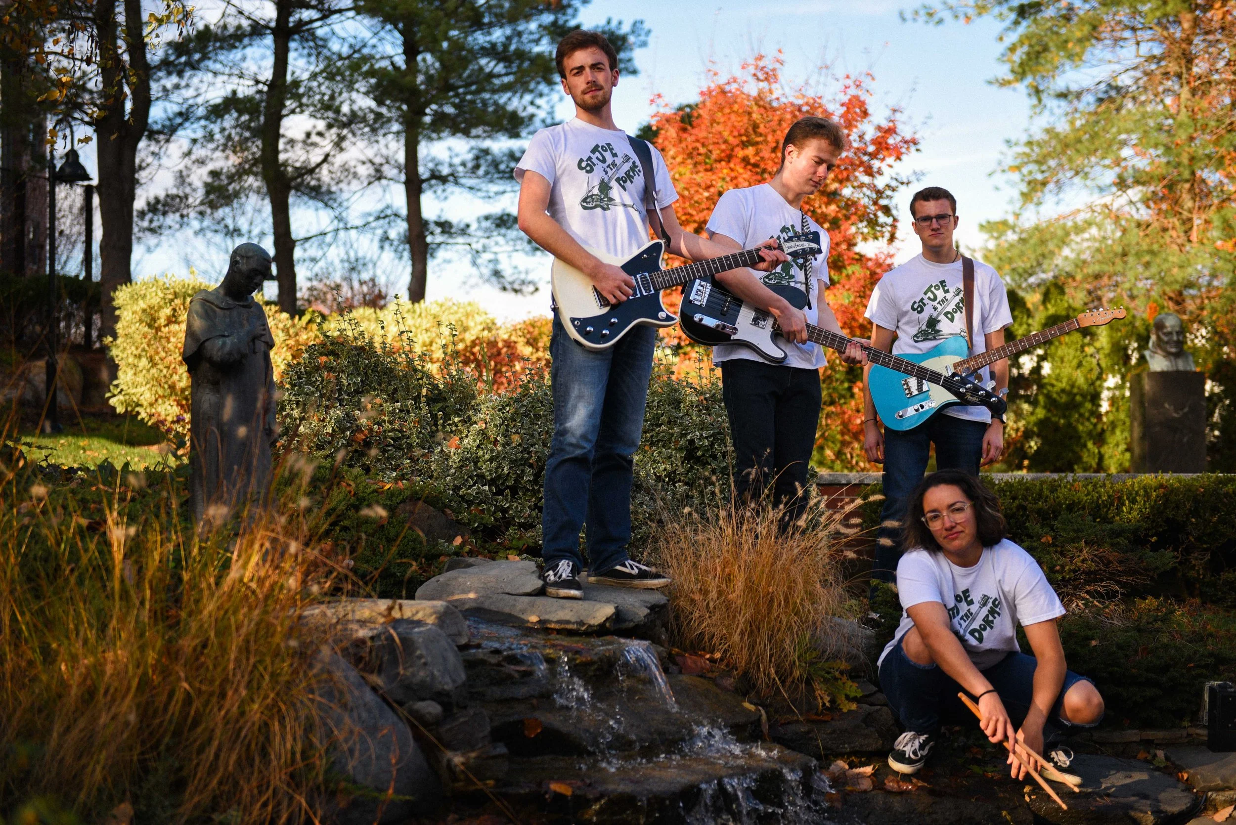 Group of five young people outdoors, four standing with guitars and one sitting on rocks, during autumn with colorful trees in background and small waterfall in foreground.
