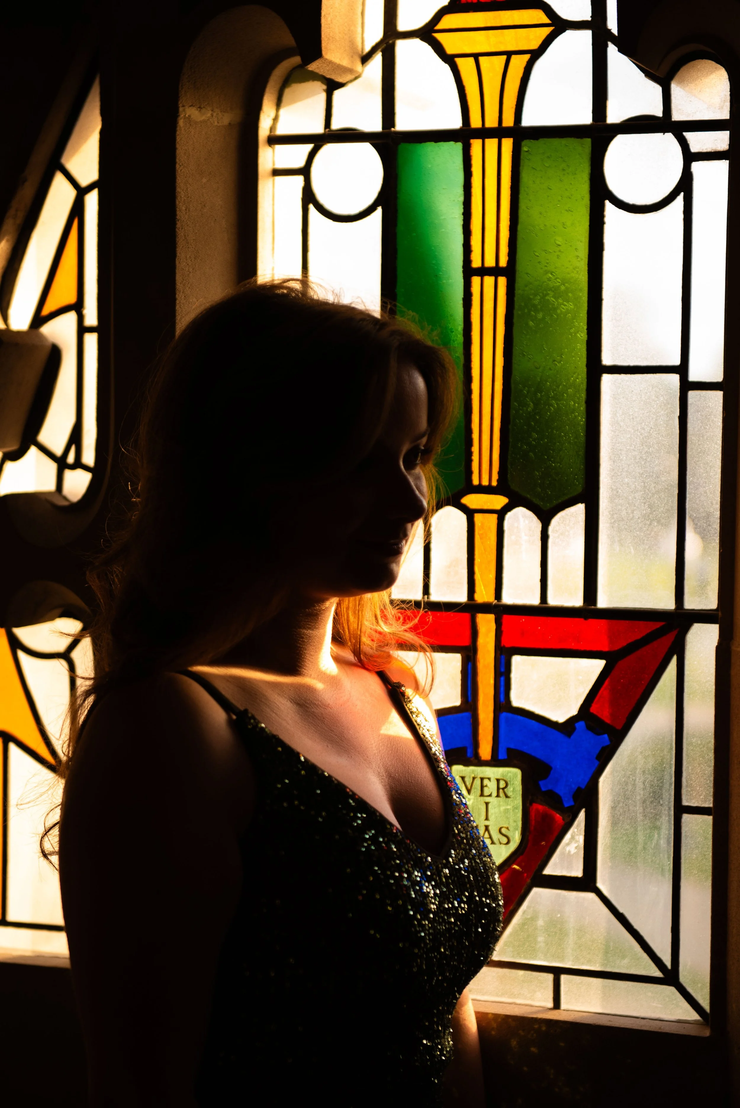 A woman with long hair standing in front of a stained glass window, with light illuminating her silhouette.