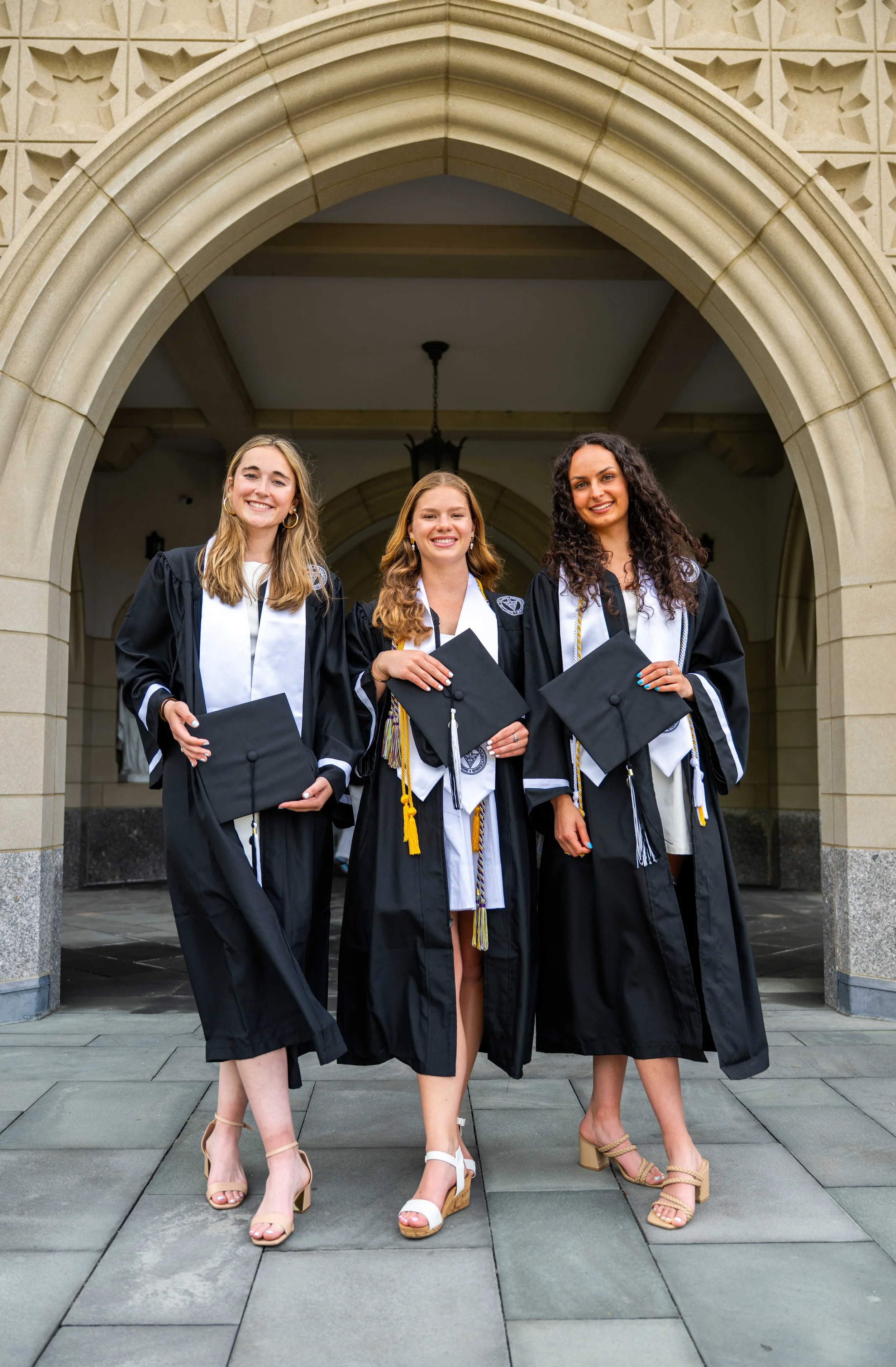 Three young women in graduation gowns and caps standing under an arched stone entryway, smiling and holding their diplomas.