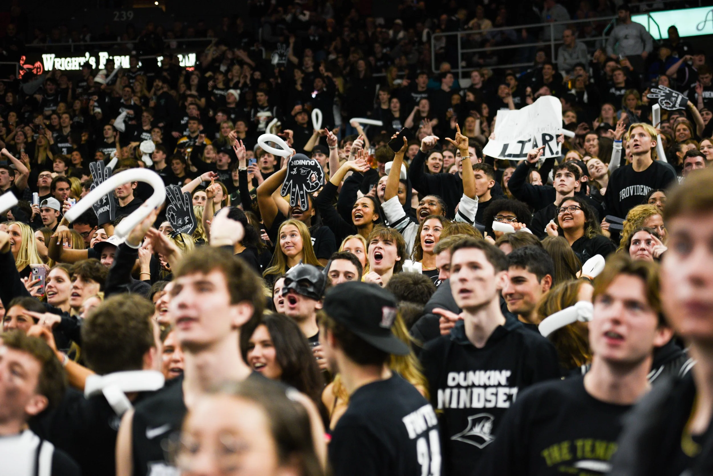 Crowd of excited young people at a sporting event, many wearing black and white clothing, cheering, holding signs, and inflatable objects in a stadium.