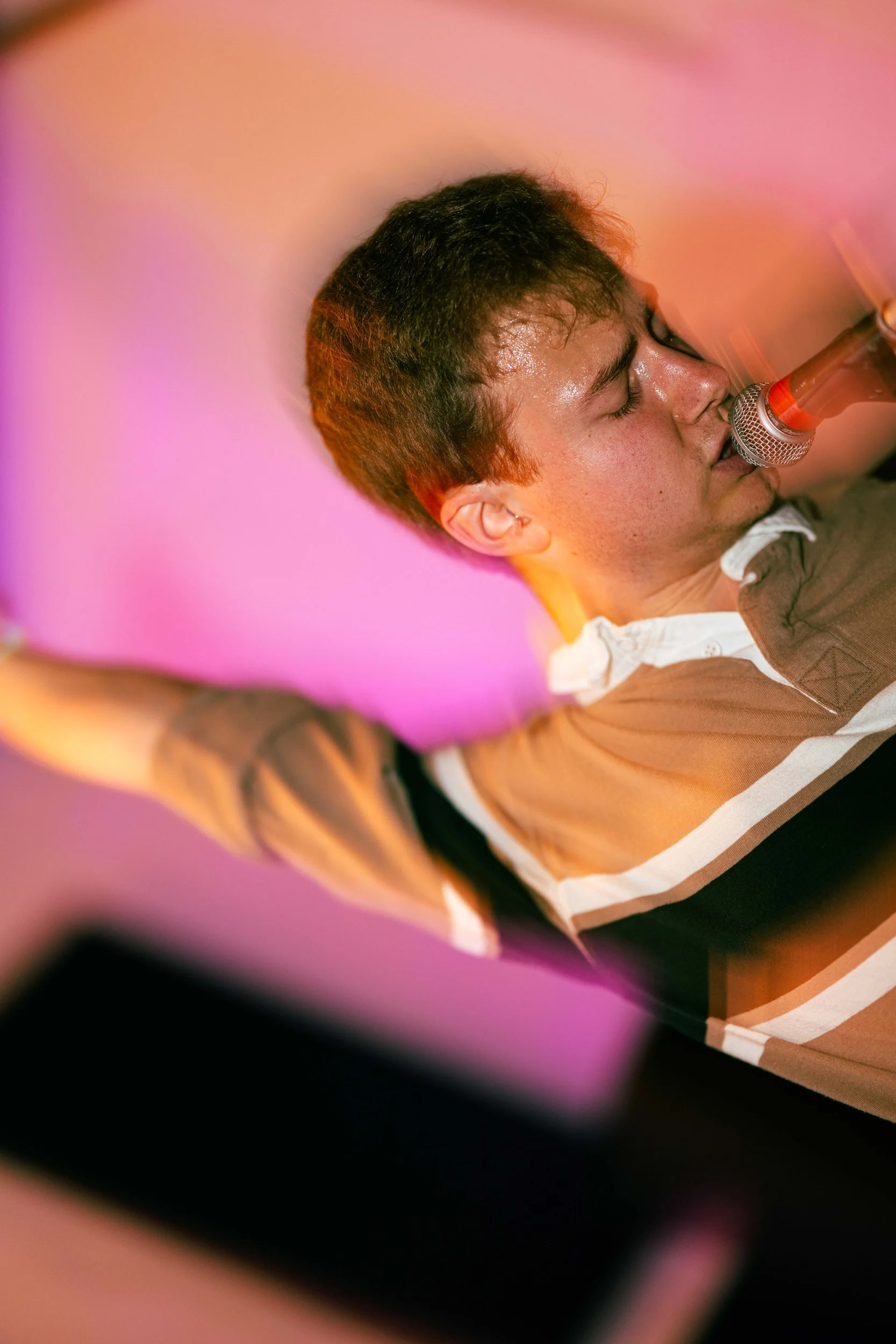 A young person with short dark hair and a tan shirt singing into a microphone with closed eyes, sweat on their face, against a pink background.