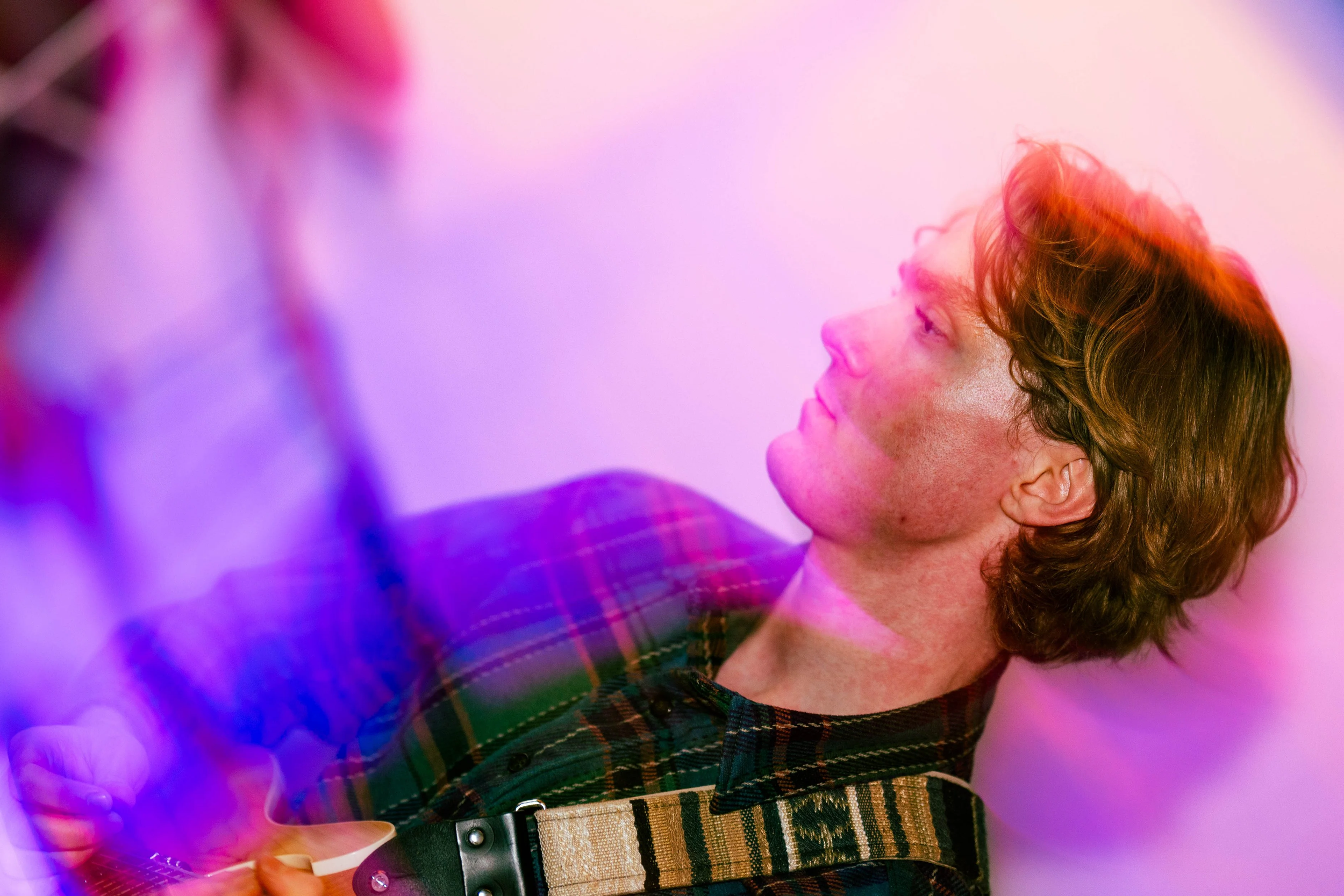 A person with wavy brown hair and freckles, wearing a plaid shirt and guitar strap, looking to the side in colorful lighting.