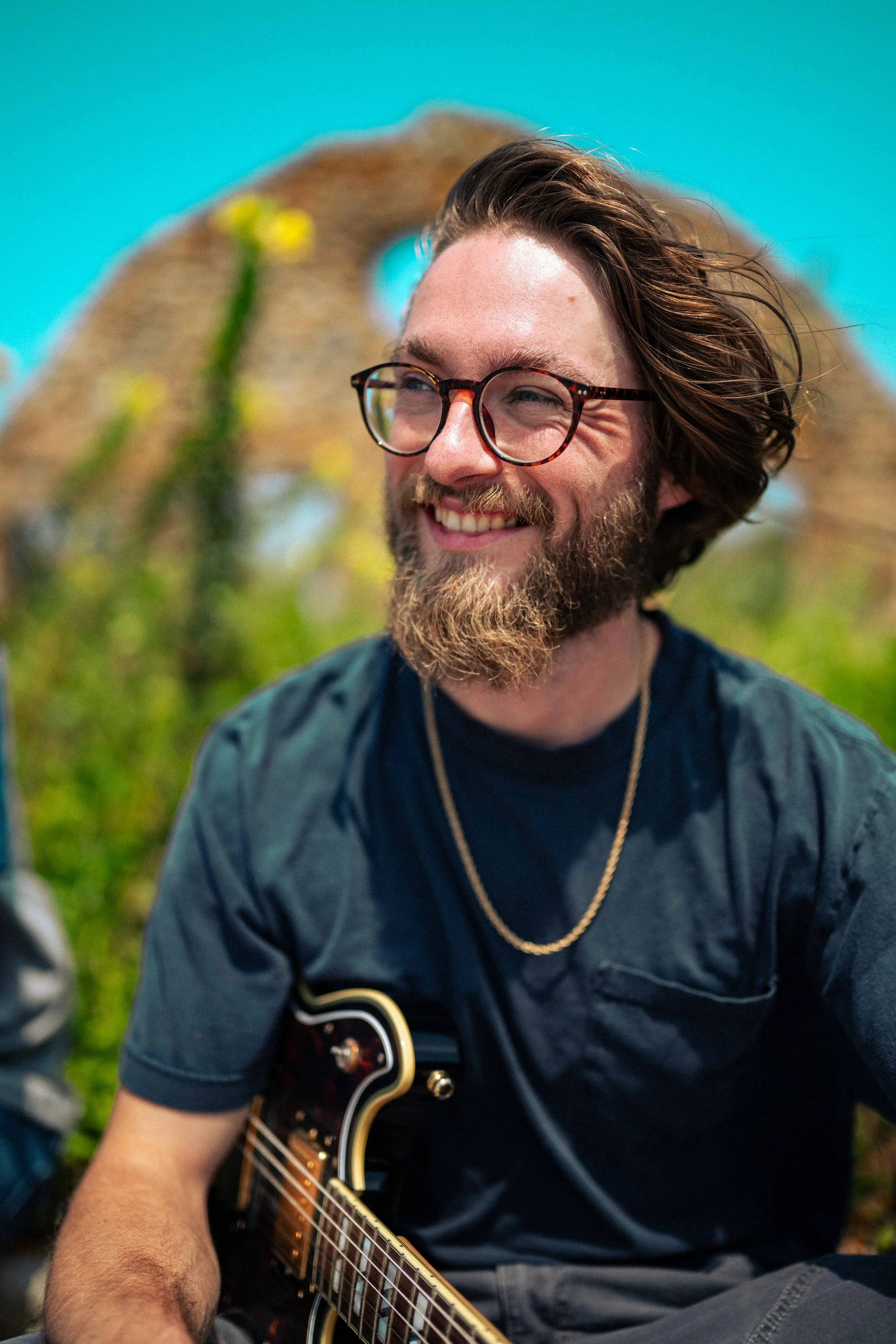 A man with glasses, a beard, and long hair smiling while holding a guitar outdoors, with a large rock formation in the background.