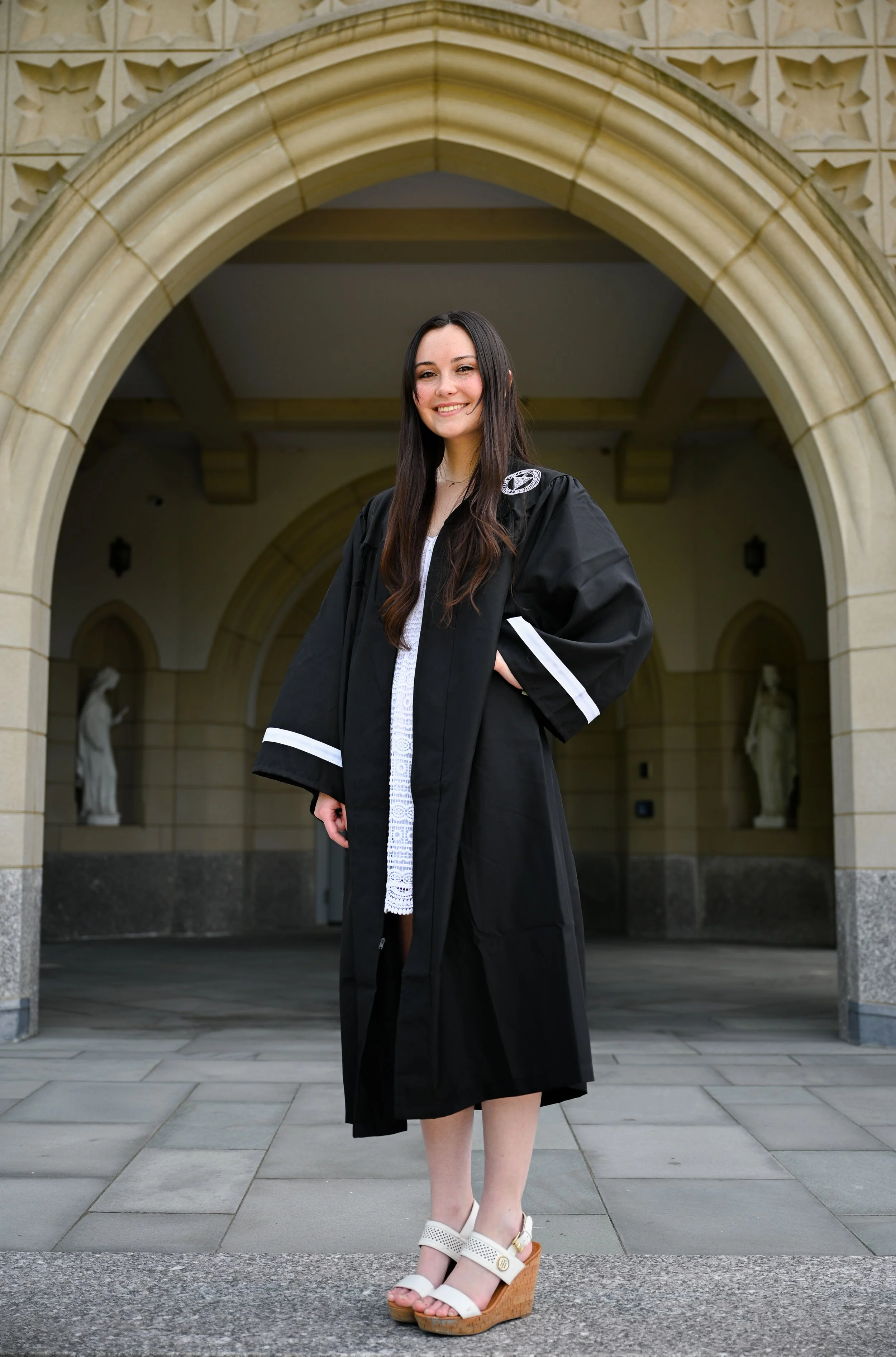 A young woman in a graduation cap and gown standing under an arched stone entrance of a historic building, smiling with her hand on her hip.