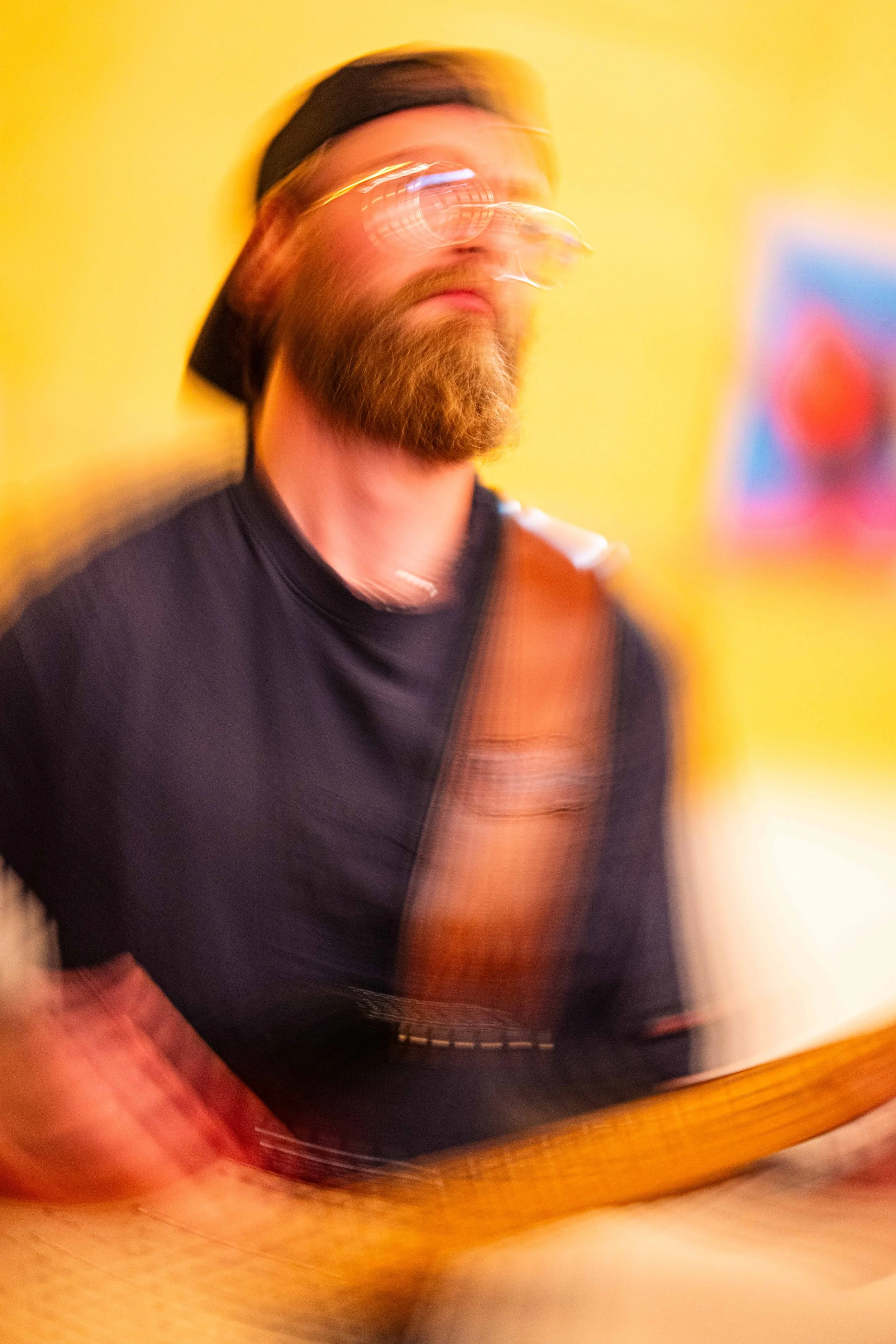 A man with a beard wearing glasses and a black hat, playing the guitar against a yellow background, with a blurred motion effect.