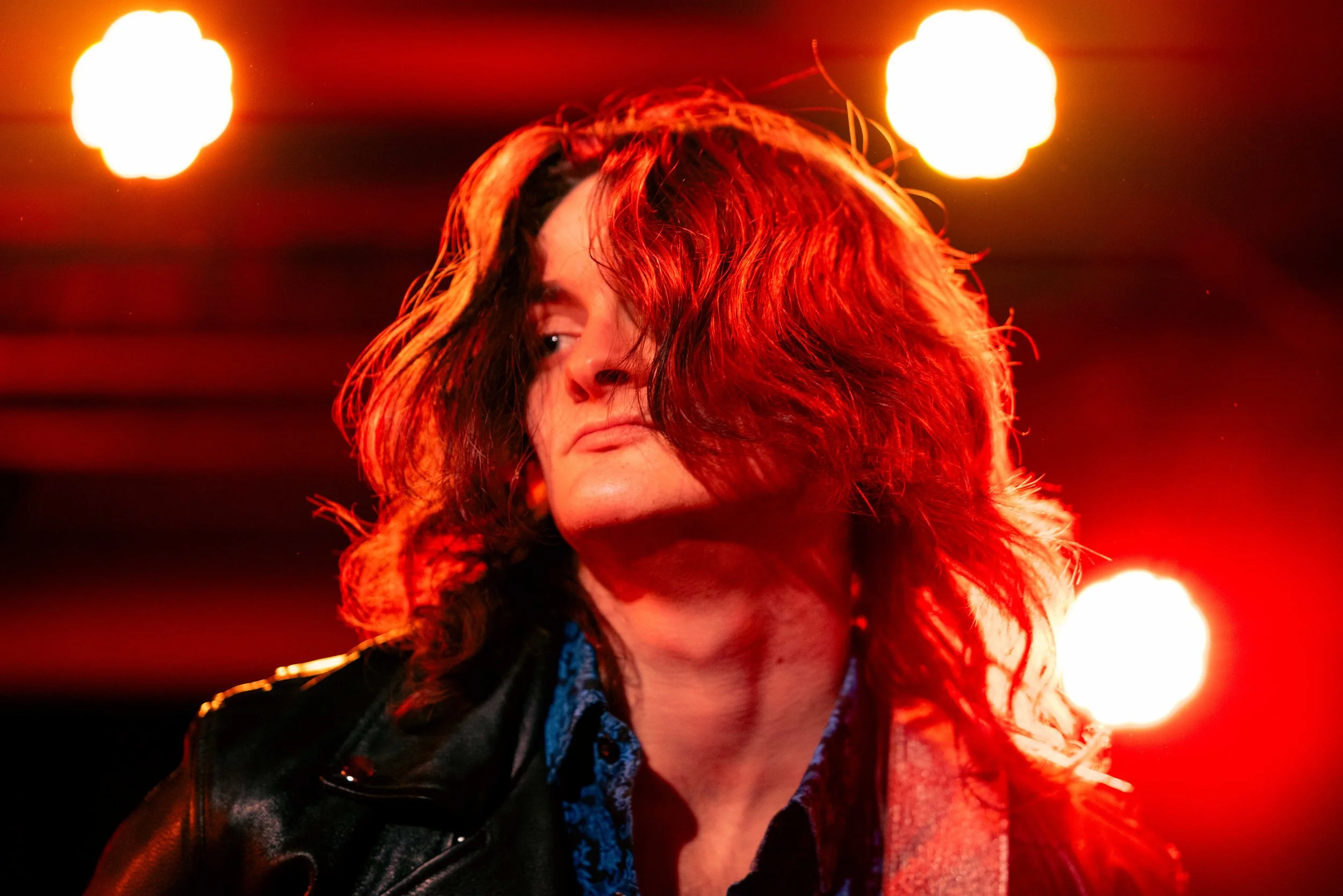 A man with long, wavy hair partially covering his face, wearing a leather jacket and a patterned shirt, standing under red stage lighting.