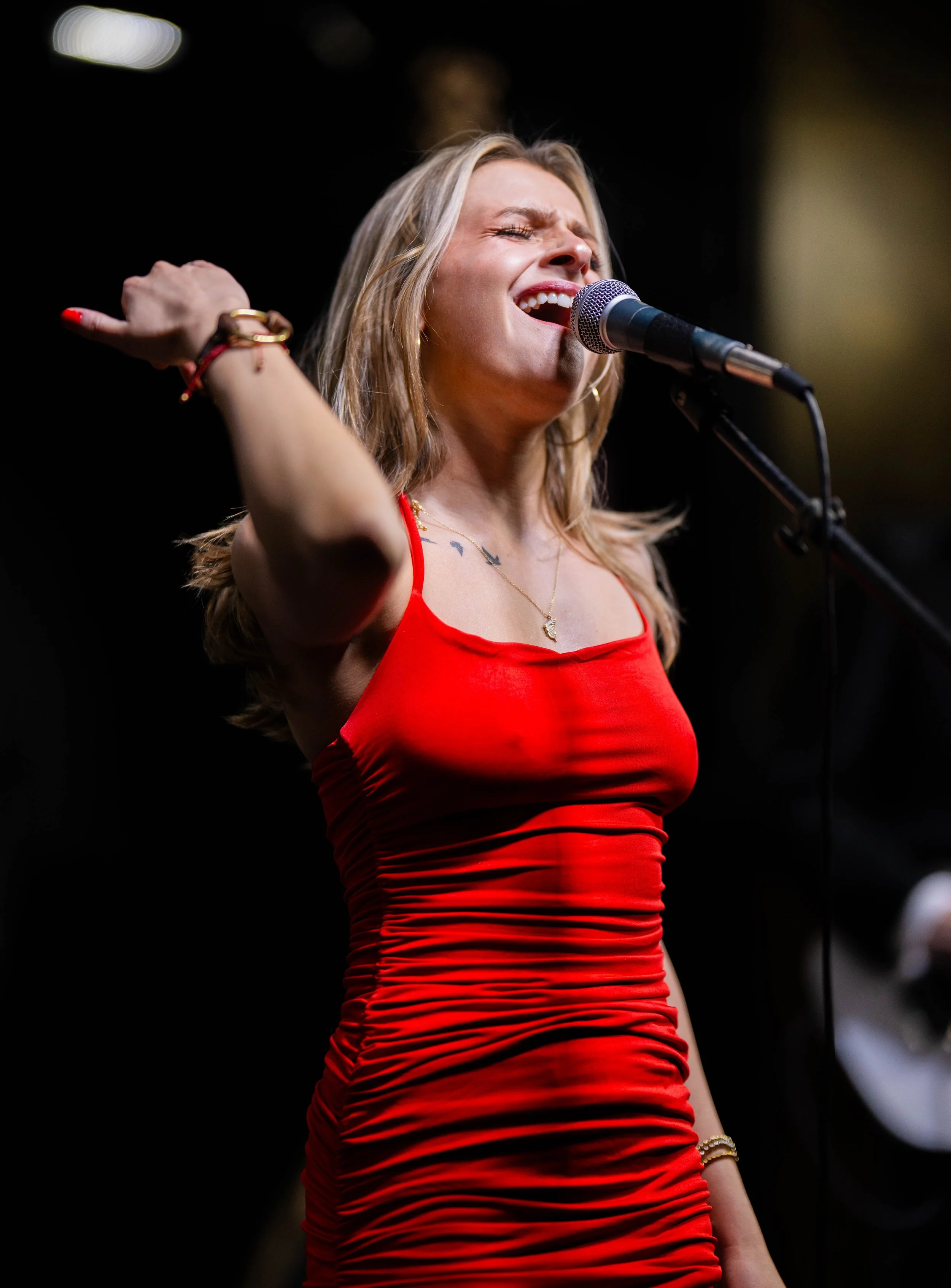 A woman singing passionately into a microphone on stage, wearing a red sleeveless dress with ruching details.