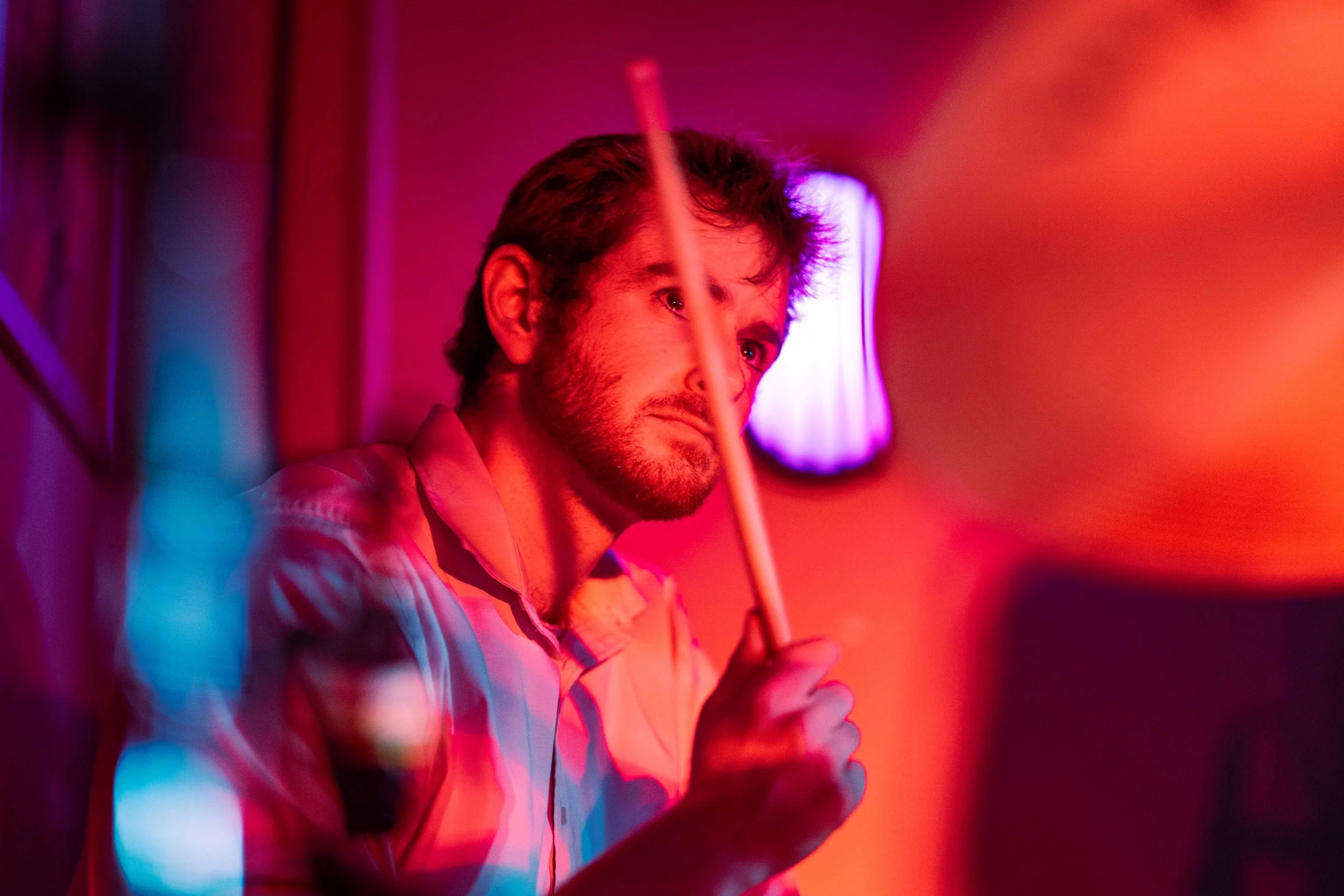 A man with brown hair and a beard is holding a drumstick, surrounded by colorful pink and purple neon lights, in a dimly lit room.
