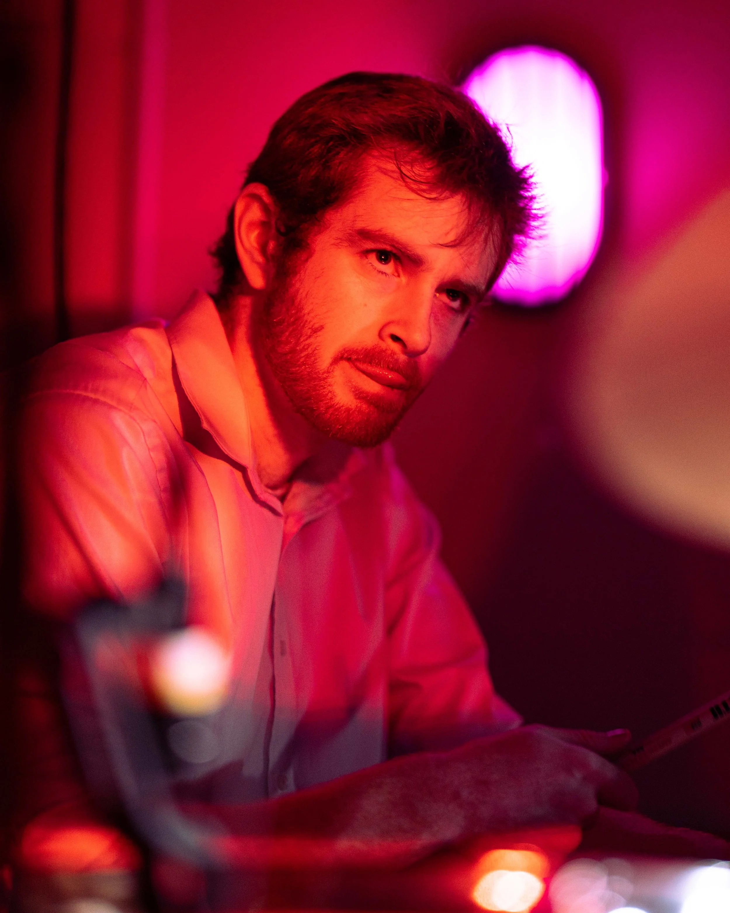 A man with dark hair and a beard, wearing a pink shirt, sitting in a dimly lit room illuminated by pink and purple neon lights, looking at the camera.