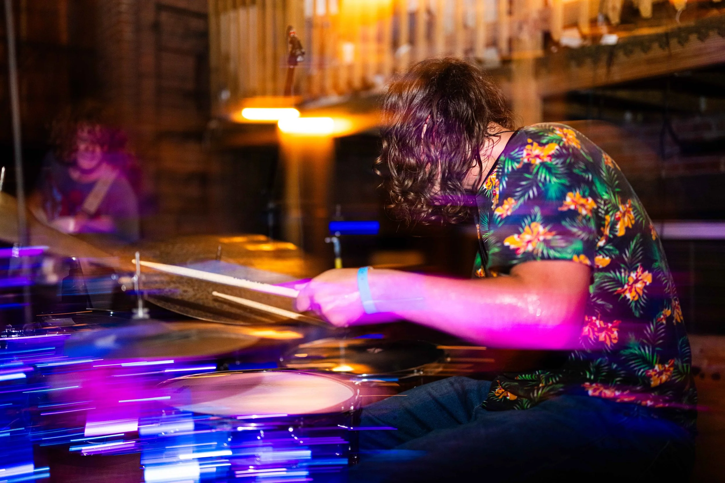 A person playing a piano in a bar or music venue with colorful lights and another person in the background. The person in the foreground is wearing a floral shirt and has curly hair.