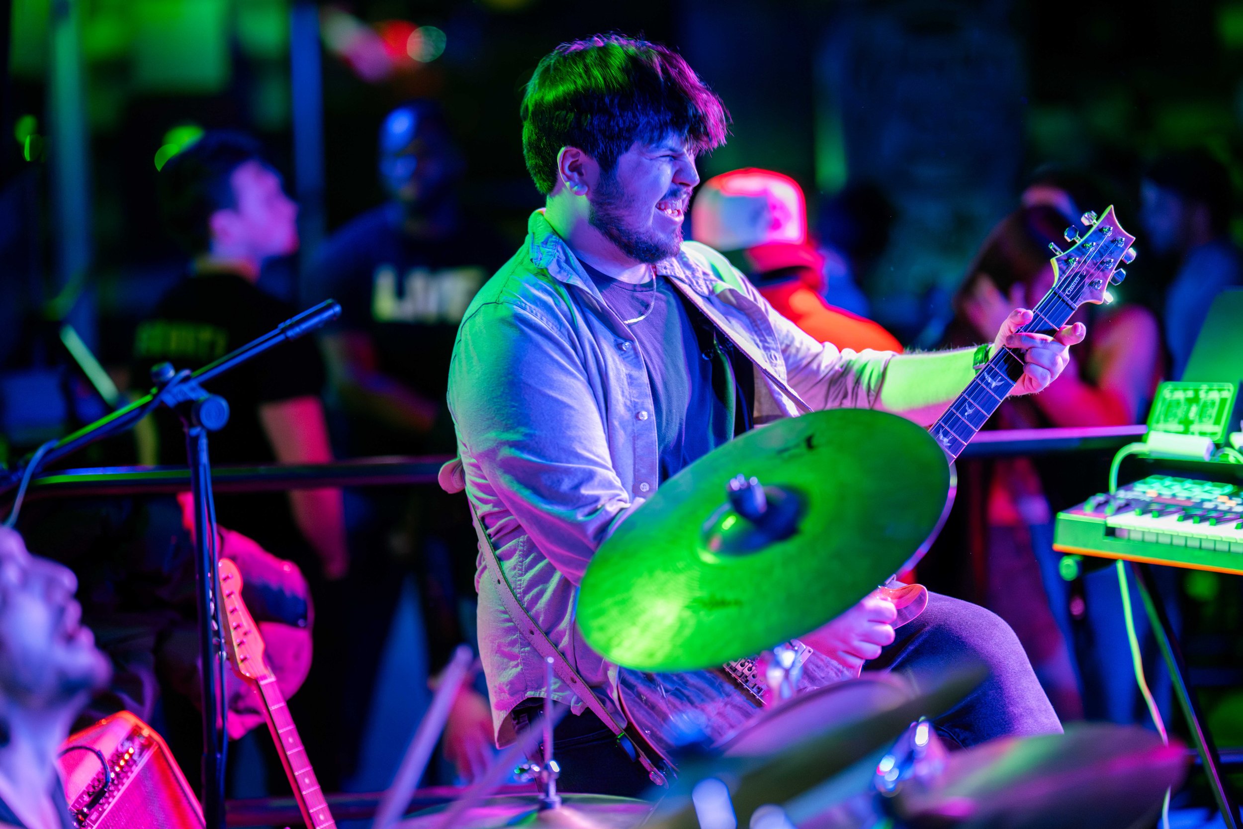 A young man playing the guitar and drums at an outdoor music event at night, with colorful stage lighting and a crowd in the background.