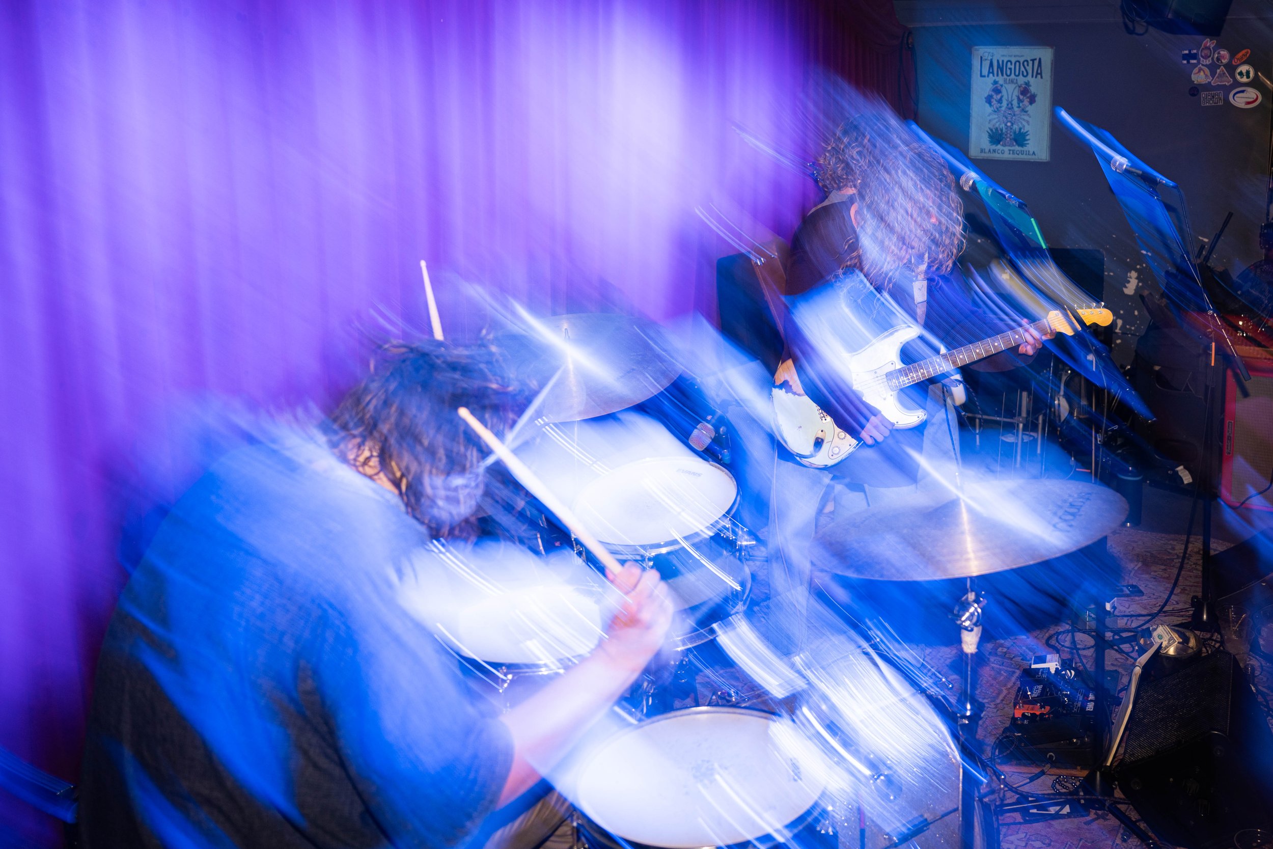 Blurred motion image of two musicians playing electric guitar and drums during a performance with purple stage lighting.