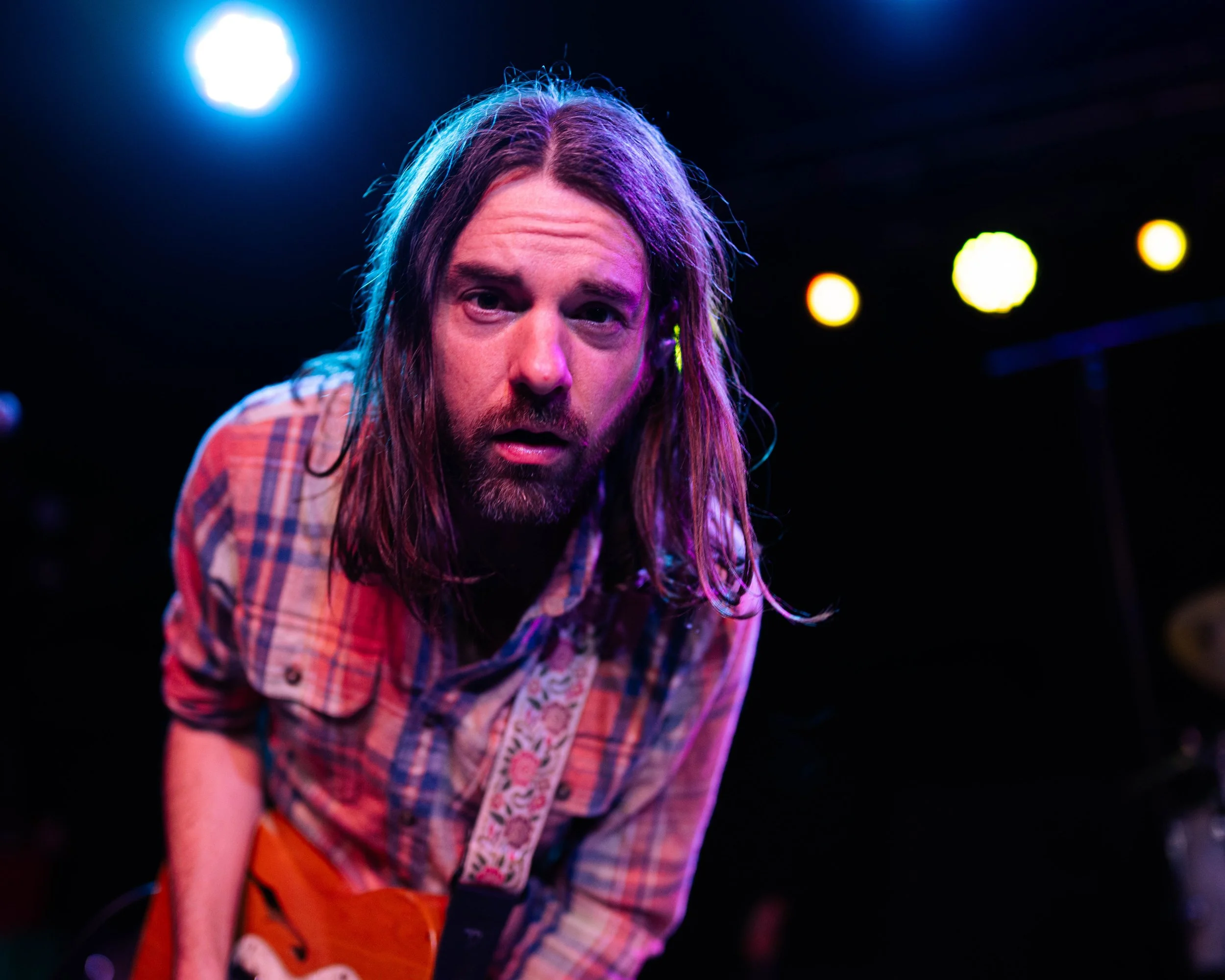 A male musician with long hair and a beard performing on stage with colorful lights.