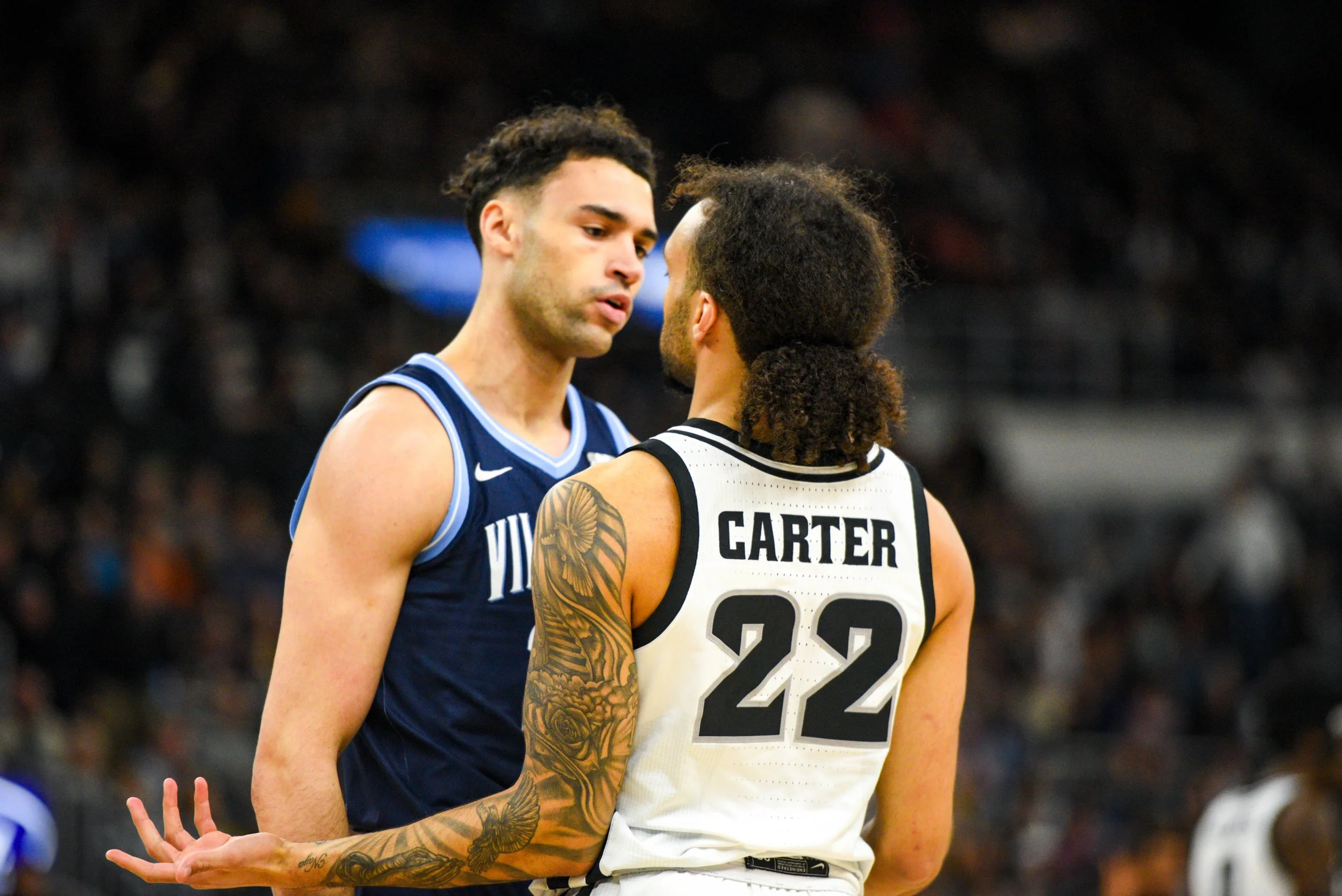 Two basketball players from opposing teams speak closely during a game; one wearing a blue jersey with white accents, the other in a white jersey with black accents and the name 'Carter' and number '22' on the back, one with tattoos on their arm, in 