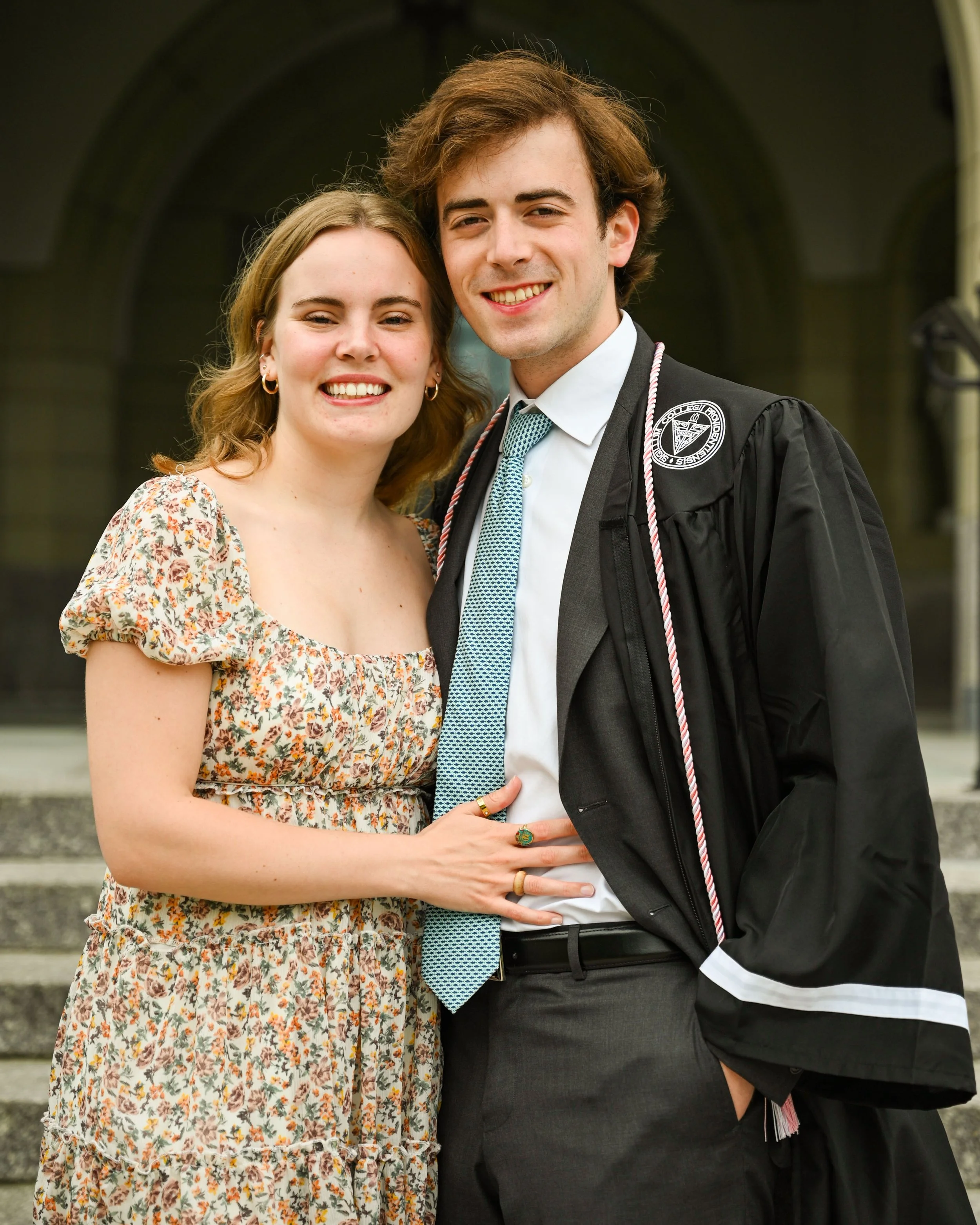 A young woman and young man smiling, standing close together in front of a building. The man is wearing a graduation gown and the woman is wearing a floral dress.
