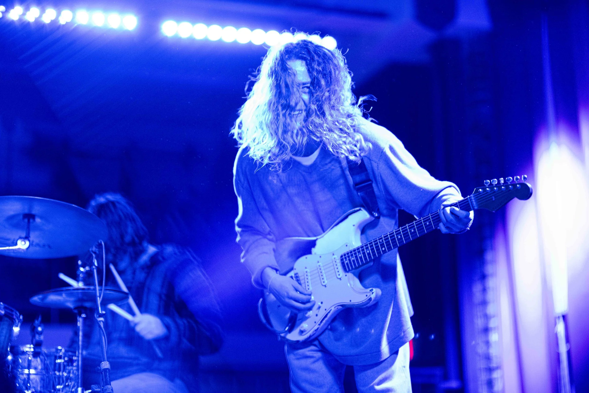 A musician with long curly hair playing an electric guitar on stage under blue lighting.
