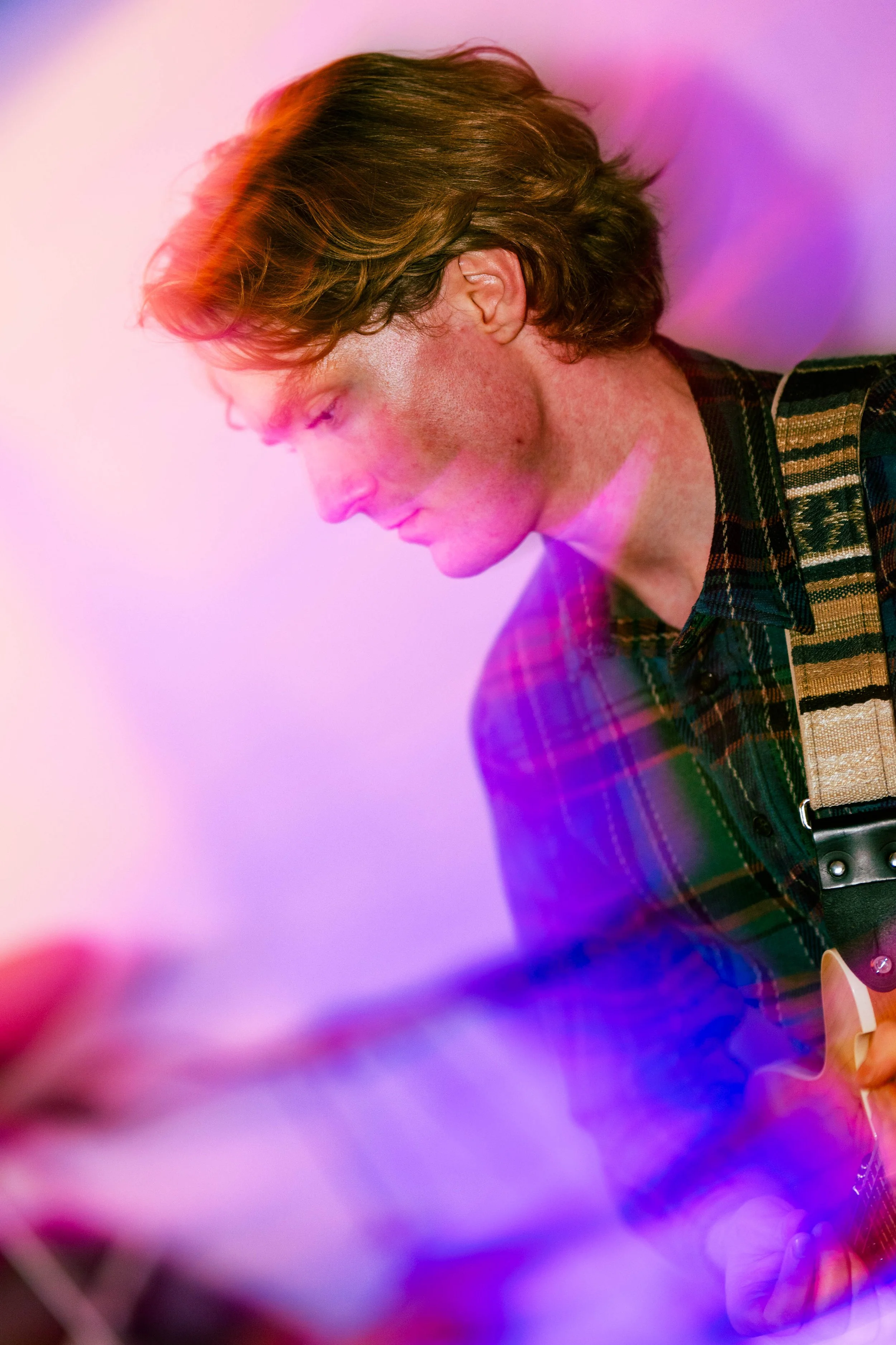 A young man with brown hair wearing a plaid shirt playing an electric guitar with colorful, artistic lighting effects.